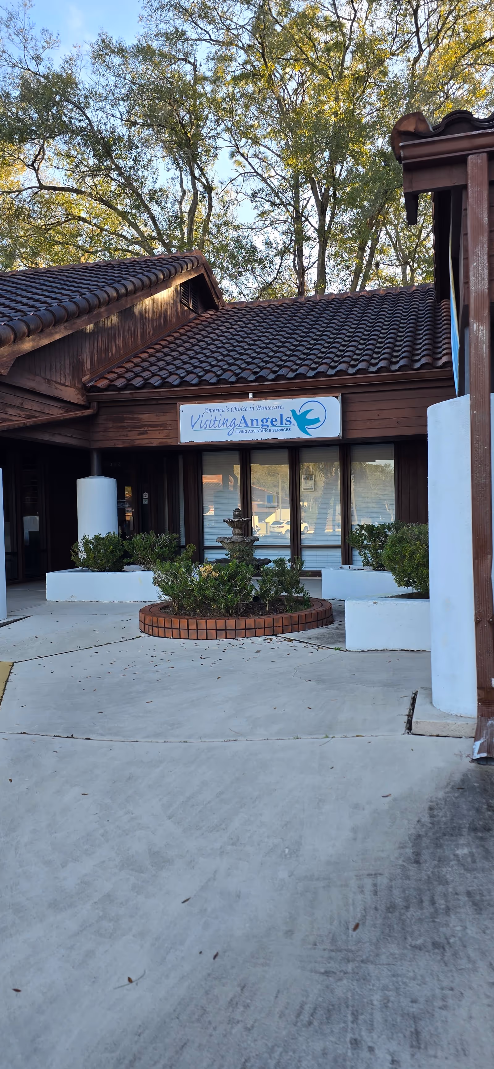 Front exterior of Visiting Angels building with sign, entryway, planters, and trees behind.