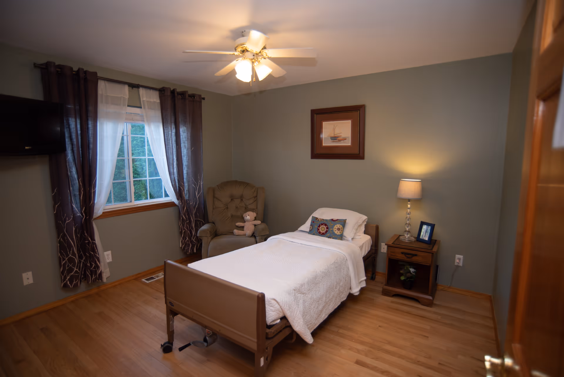 A cozy bedroom in a senior living facility with a bed, chair, nightstand, and window.