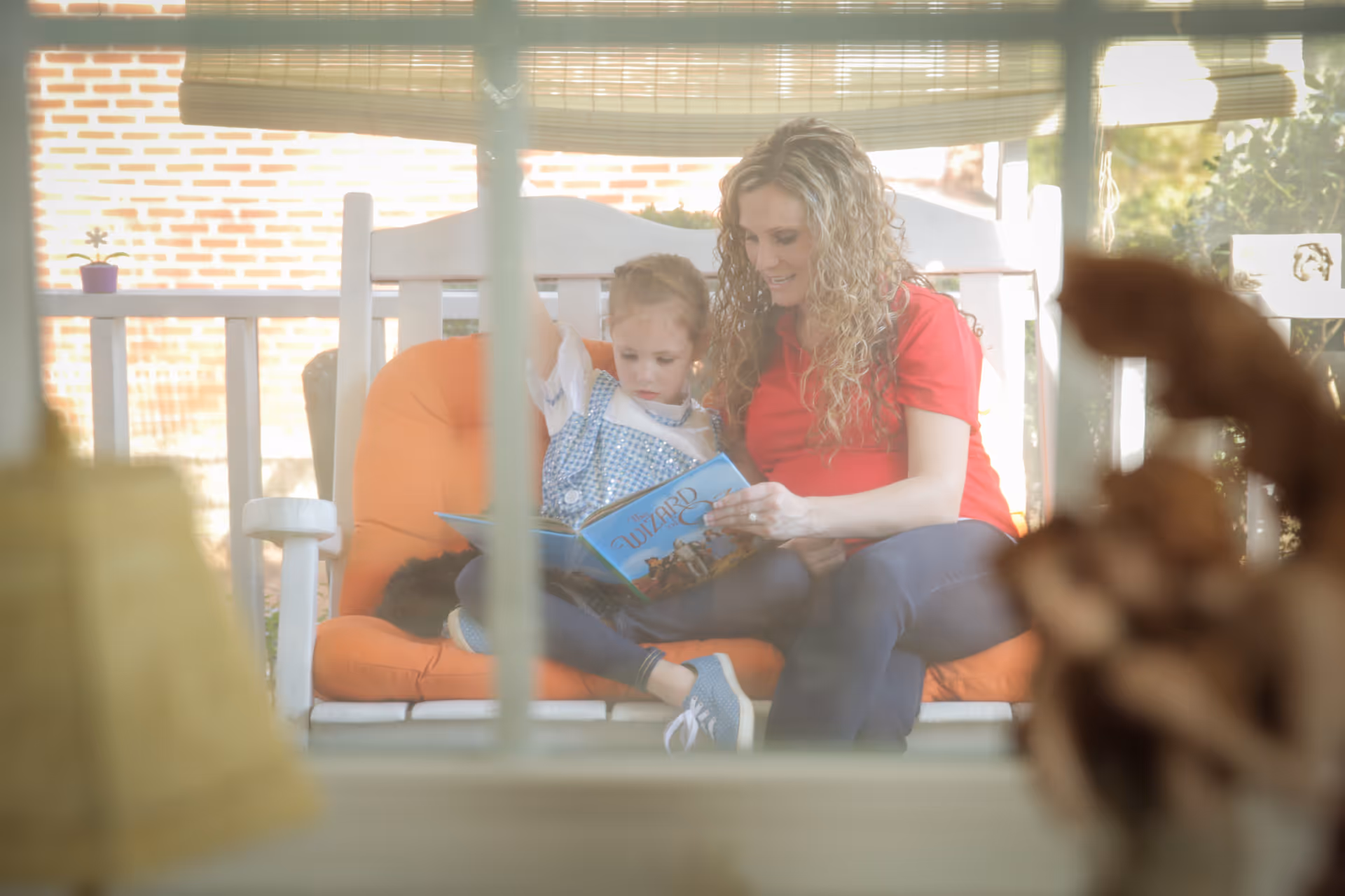 A smiling caregiver and child sit on a white porch swing reading a book outdoors.