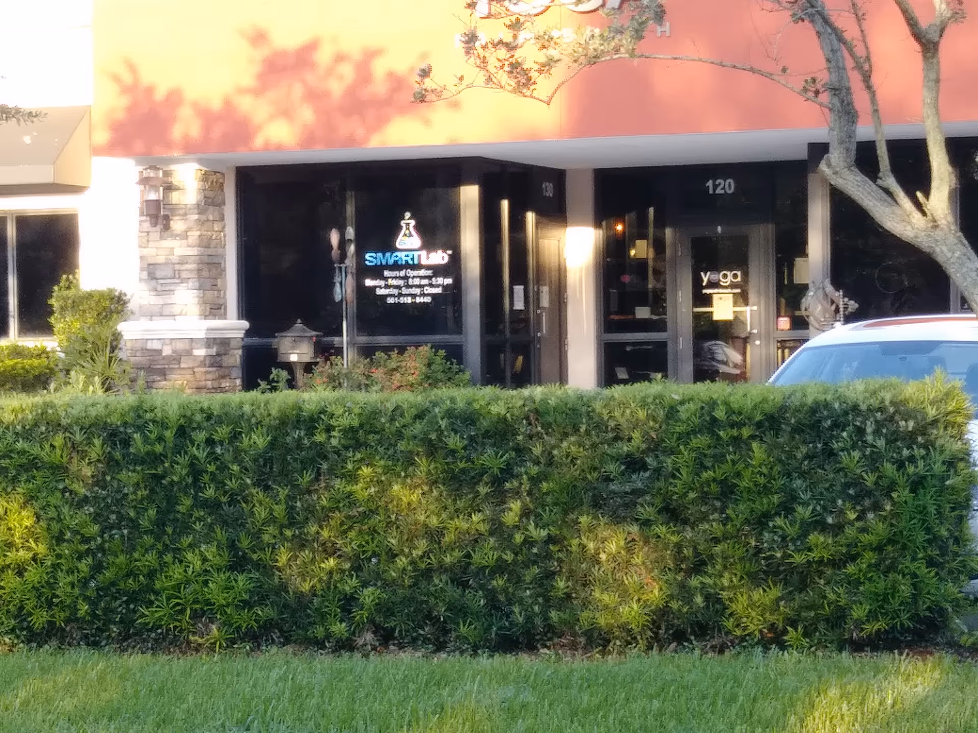 Front exterior of a storefront with glass doors and a green hedge in front.
