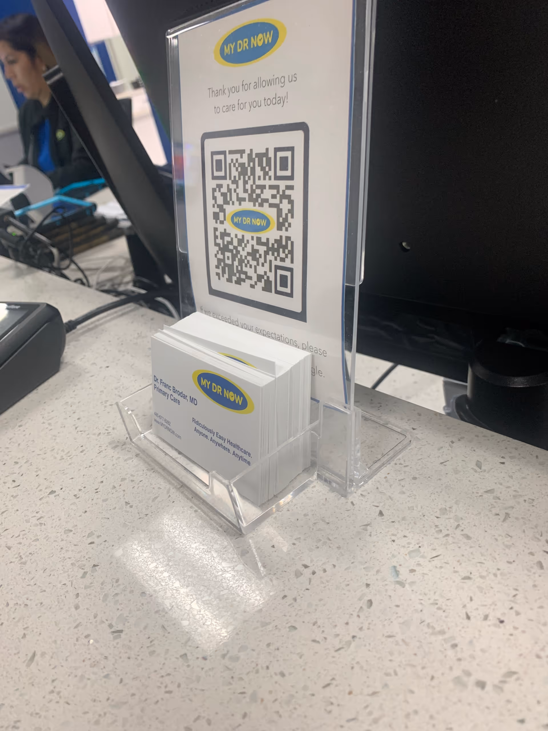 Reception counter with a My Dr Now QR code stand and a blurred staff in background.