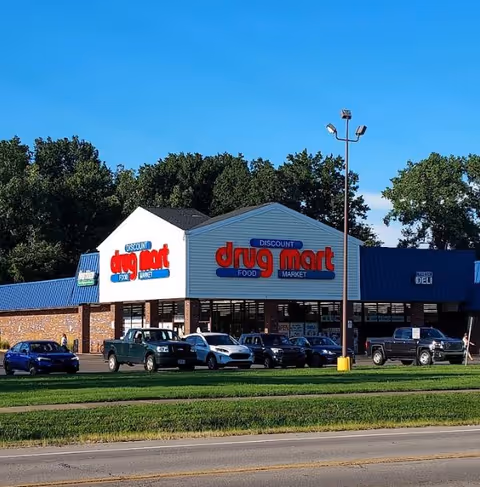 Front view of a Discount Drug Mart store with blue awning and parked cars.