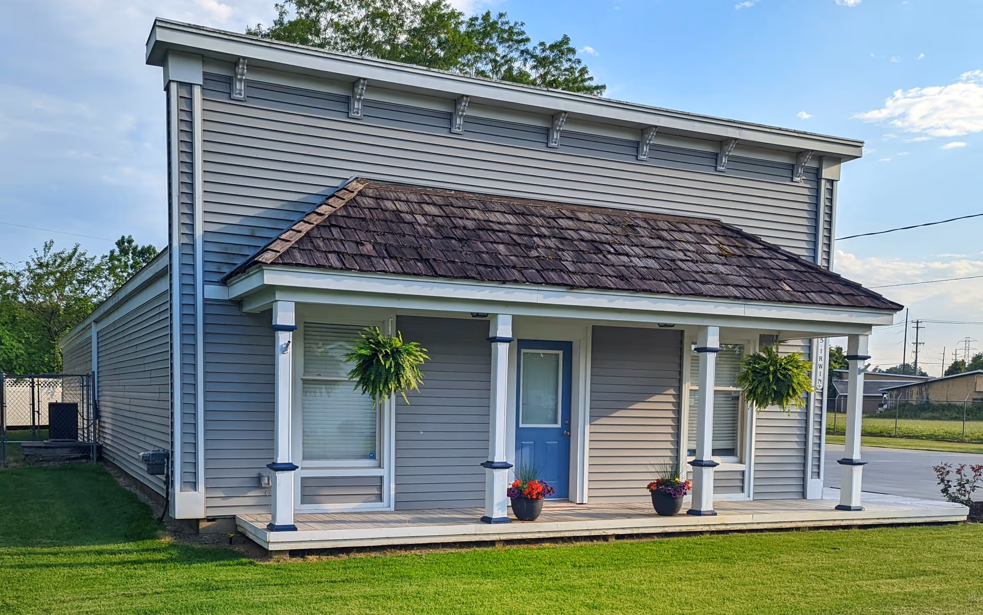 Gray building with a front porch, blue door, hanging ferns, and potted plants.