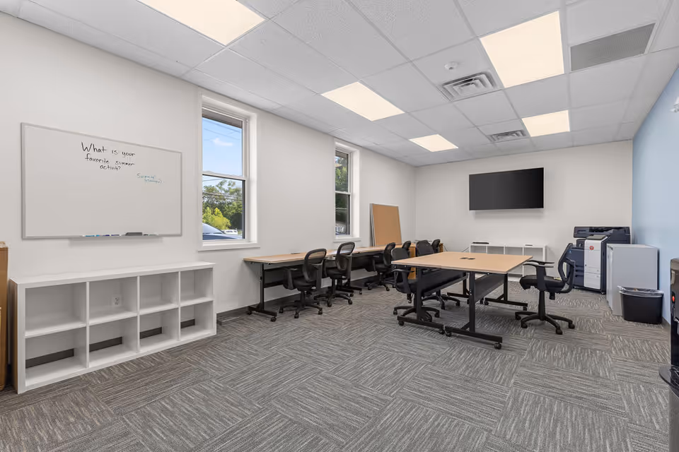 Bright classroom-style meeting room with desks, chairs, a whiteboard, and a wall-mounted screen.