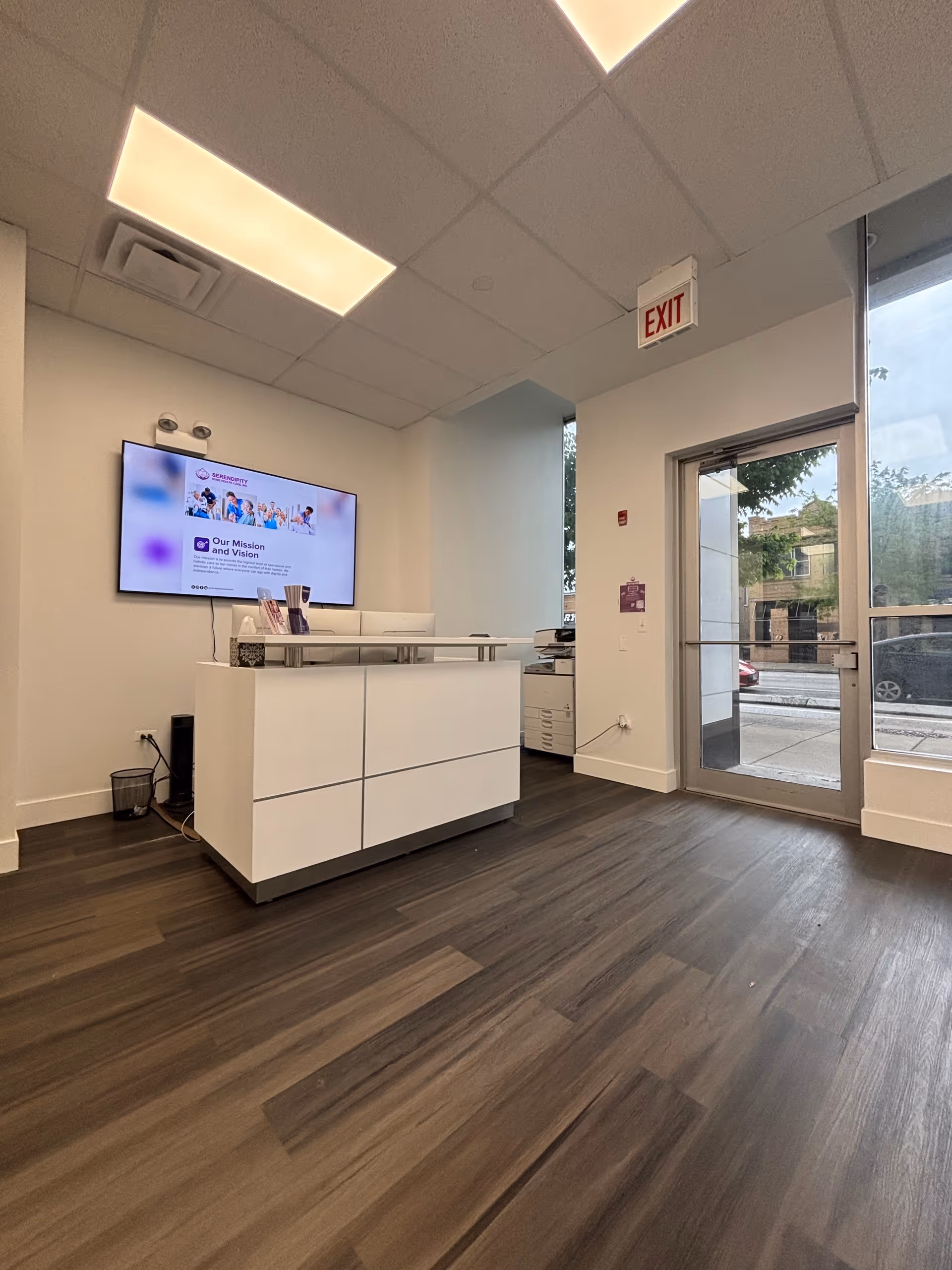 Modern reception area with white desk, wall TV, exit door, and large windowed entry.
