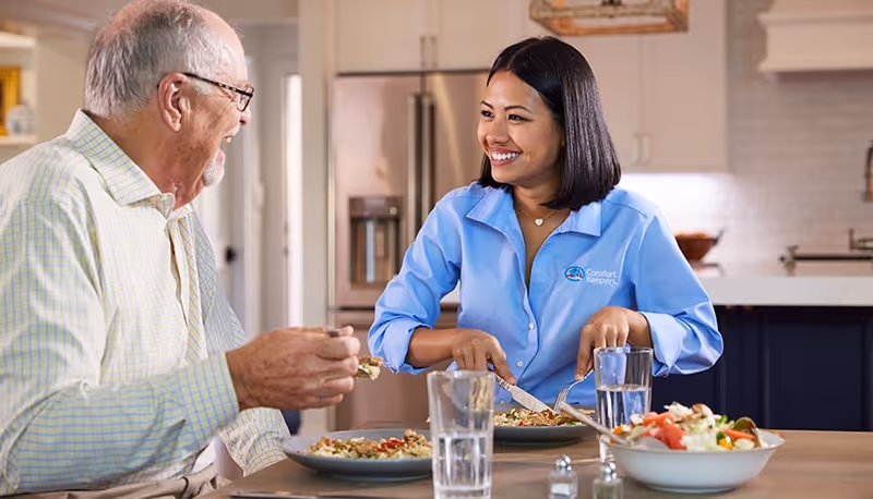 Caregiver and elderly man share a meal at a dining table in a modern kitchen.