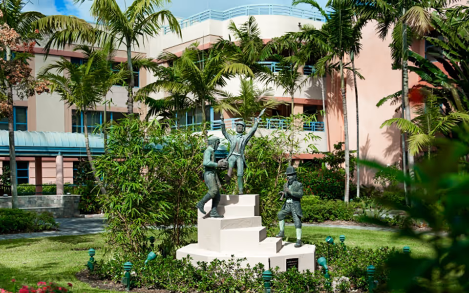 Statues atop a stepped pedestal amid a palm-filled courtyard beside a pink building.