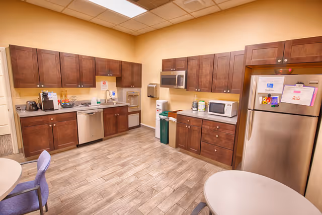 A spacious staff kitchen area with dark wood cabinets, stainless appliances, and round tables.