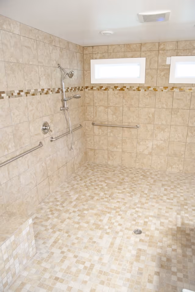 Accessible tiled bathroom with grab bars, handheld shower, and two small high windows.