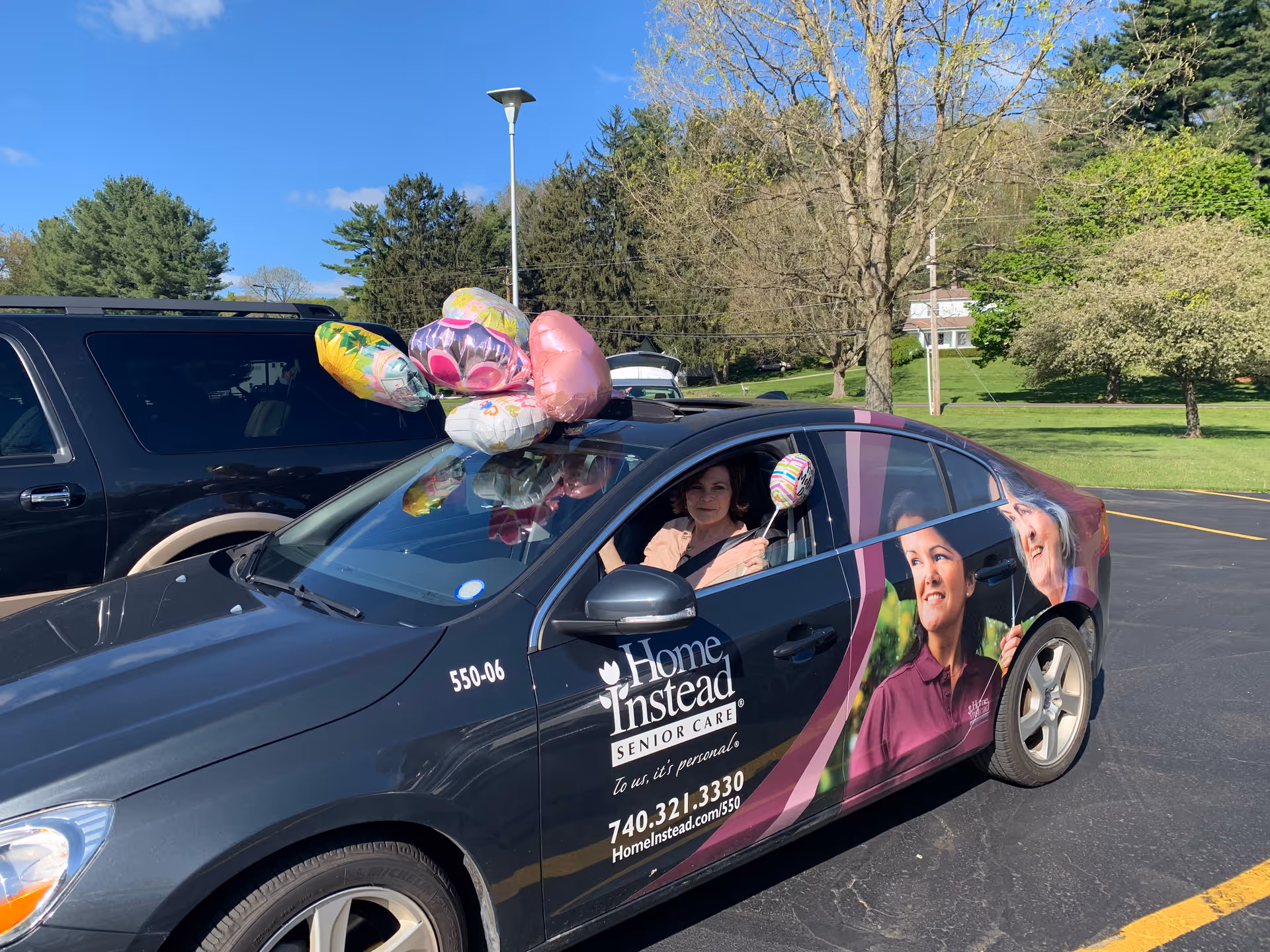 A branded Home Instead car in a parking lot with balloons and a driver smiling