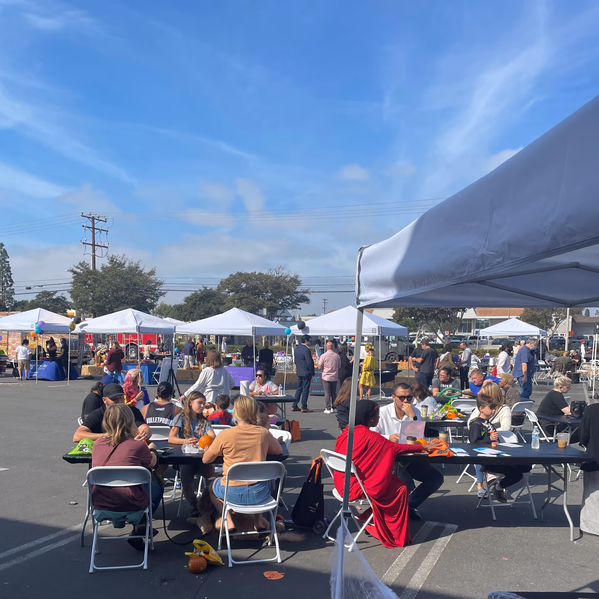 Outdoor community event with tables and white tents in a parking lot, people socializing.