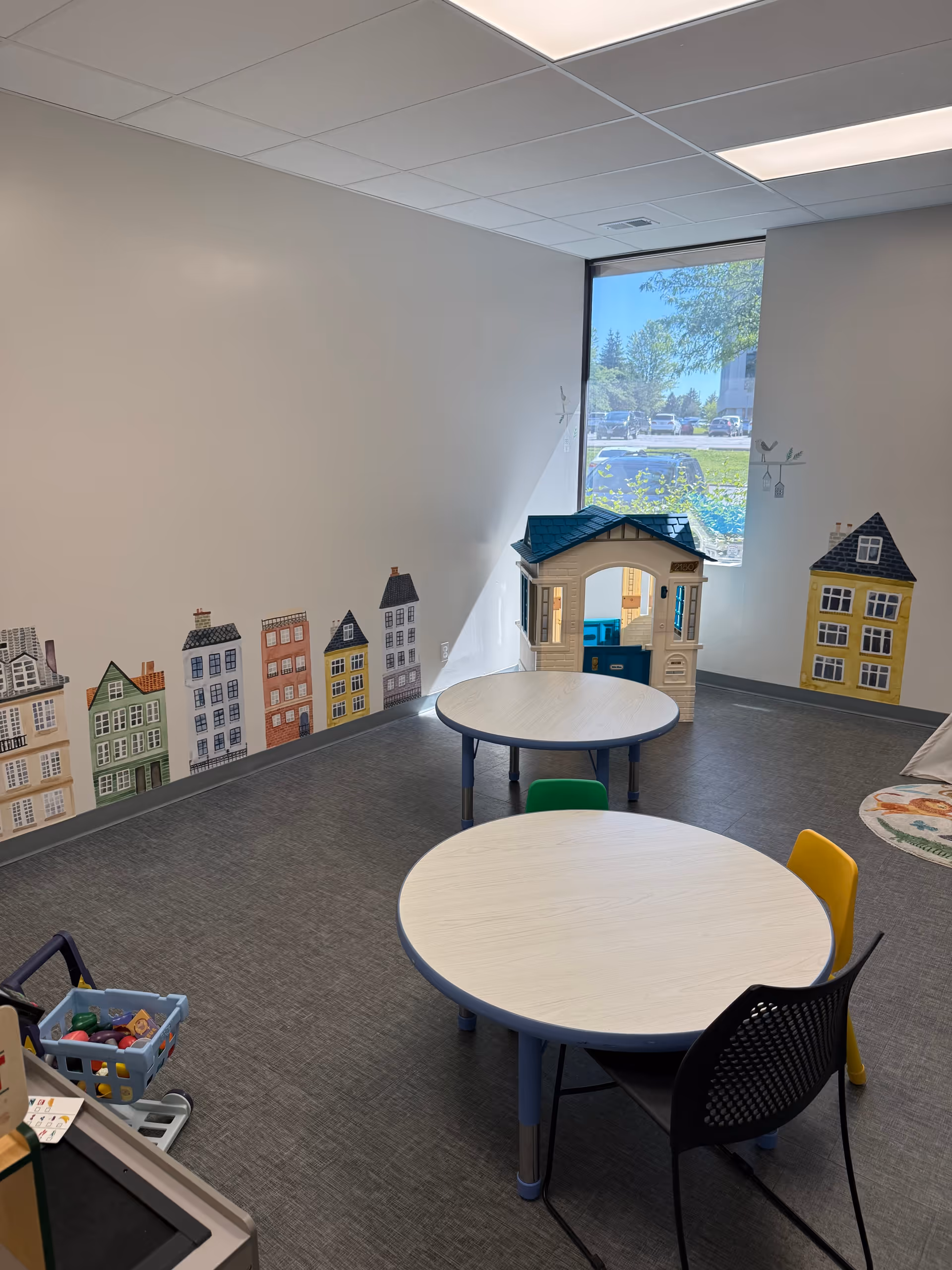 Bright indoor playroom with round tables, colorful chairs, a toy house, and wall city decals.