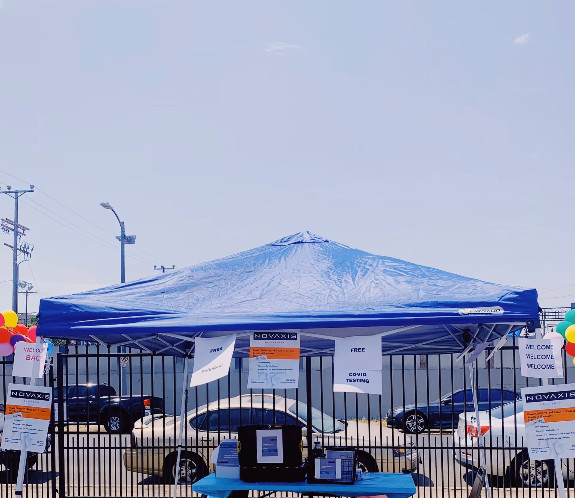 Blue canopy tent with Novaxis signage at an outdoor event behind parked cars.