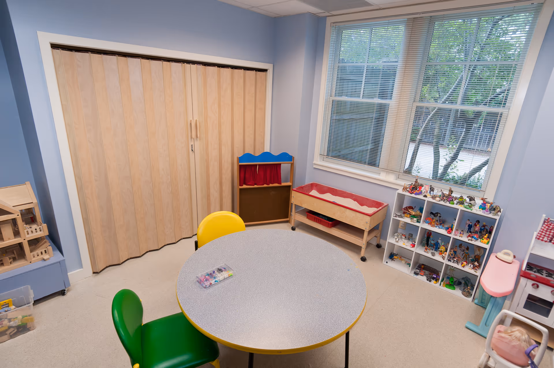 Bright children's playroom with round table, colorful chairs, and toy shelves against blue walls.