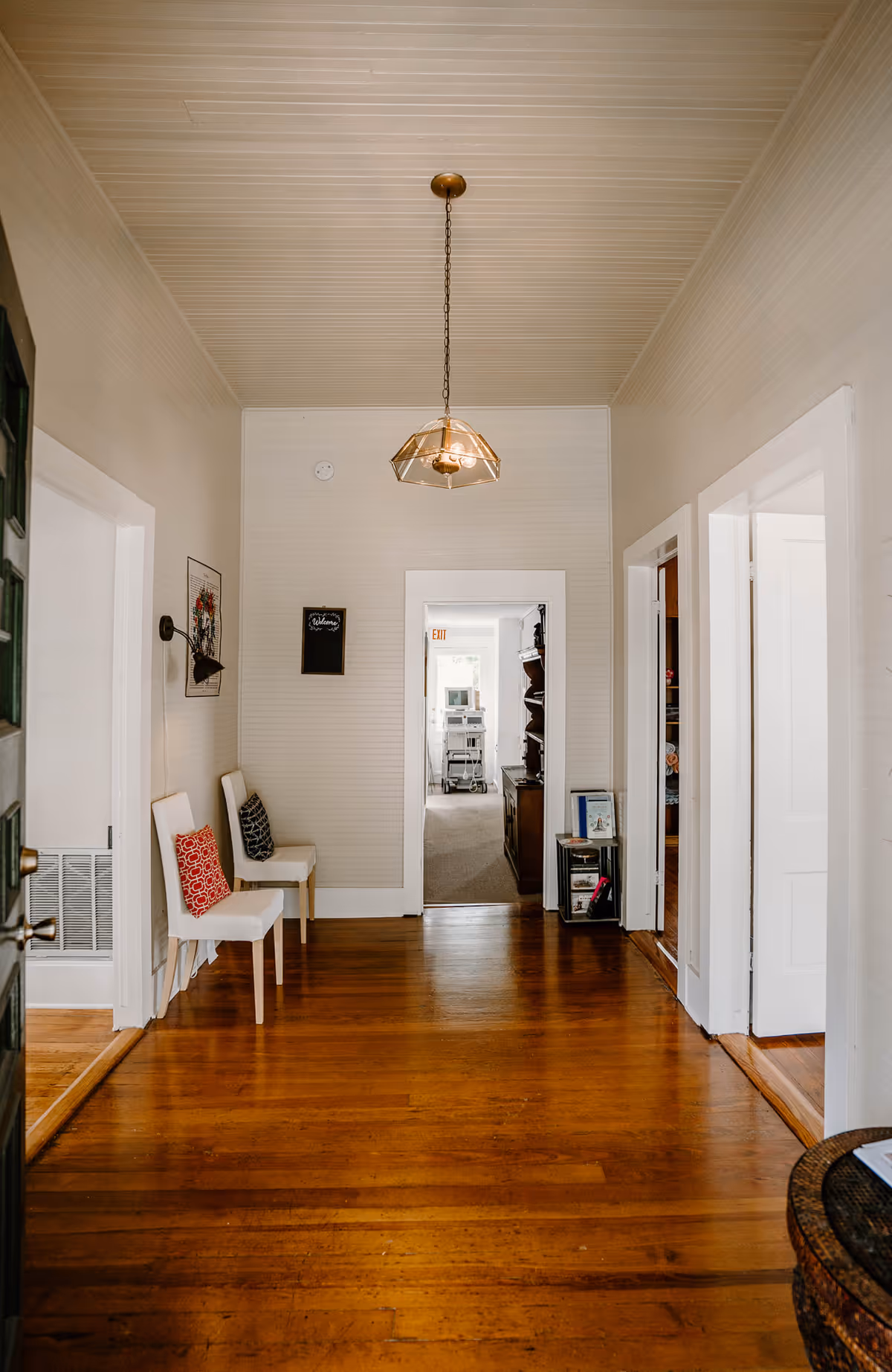 A bright hallway with wooden floors, white walls, and chairs along the left wall.