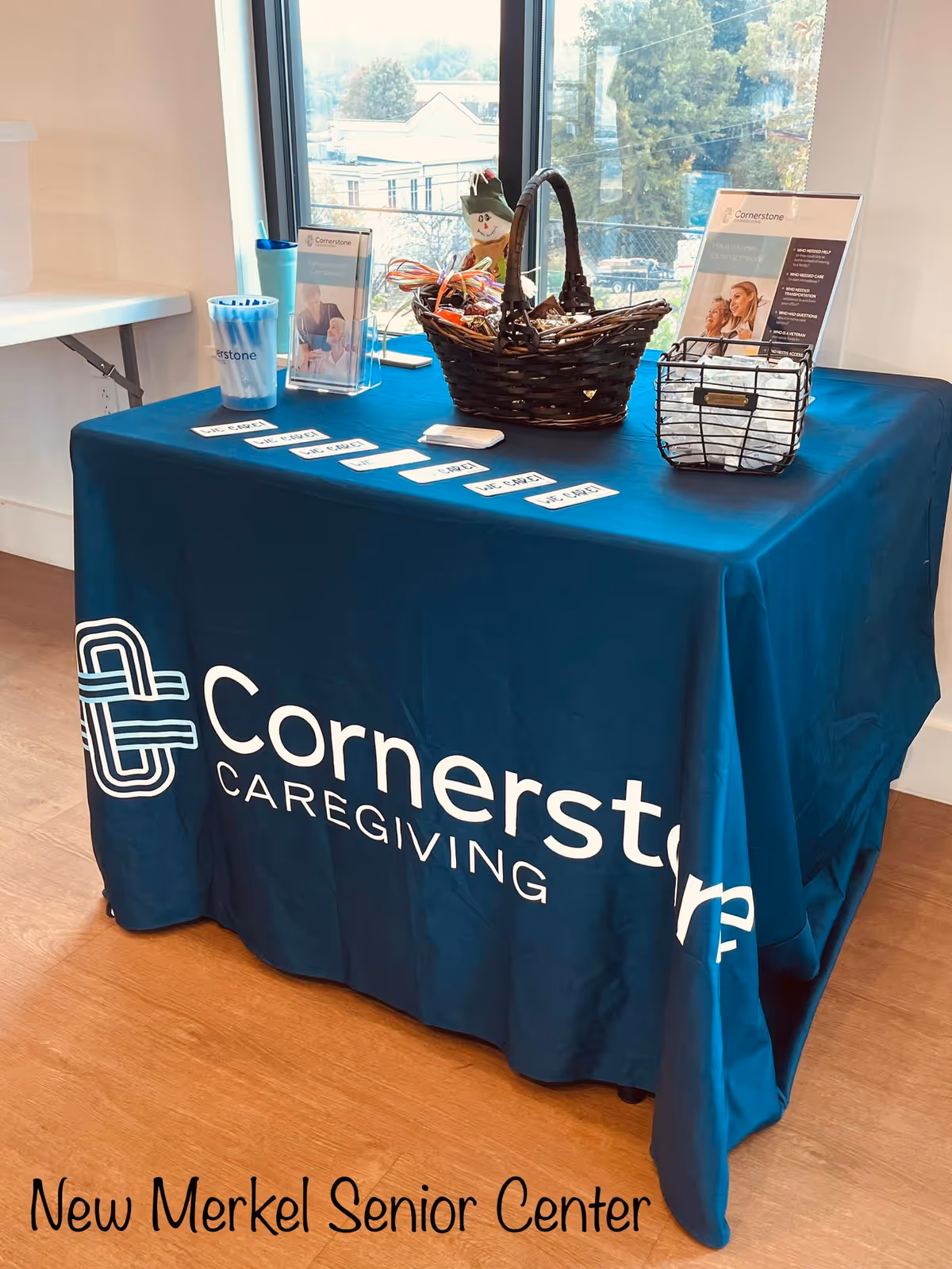 Indoor display table with blue Cornerstone Caregiving tablecloth, brochures, and a basket by a window.