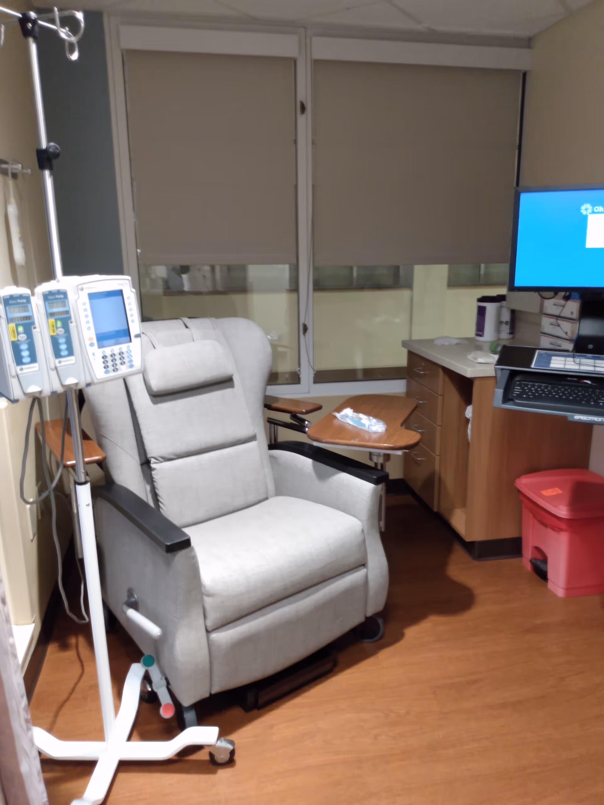 A padded infusion chair in a small clinic room with IV poles, a desk, and monitor.