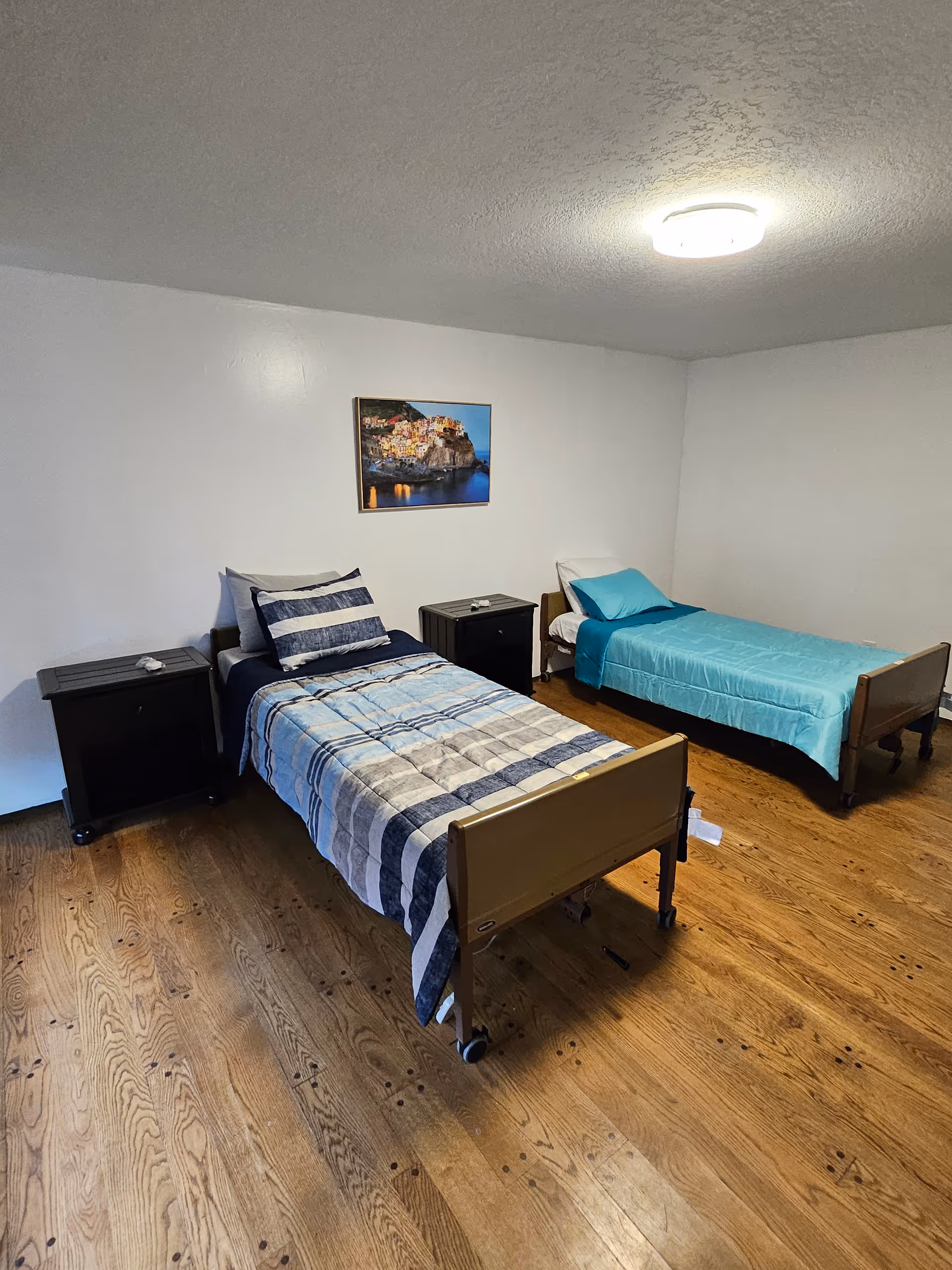 Two twin beds with nightstands and a wall painting in a simple senior living facility bedroom.