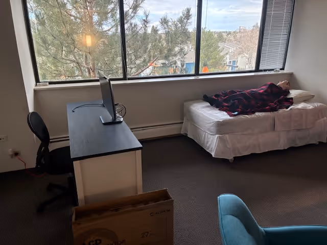 A furnished senior living bedroom with a desk, chair, and resident on the bed by window.