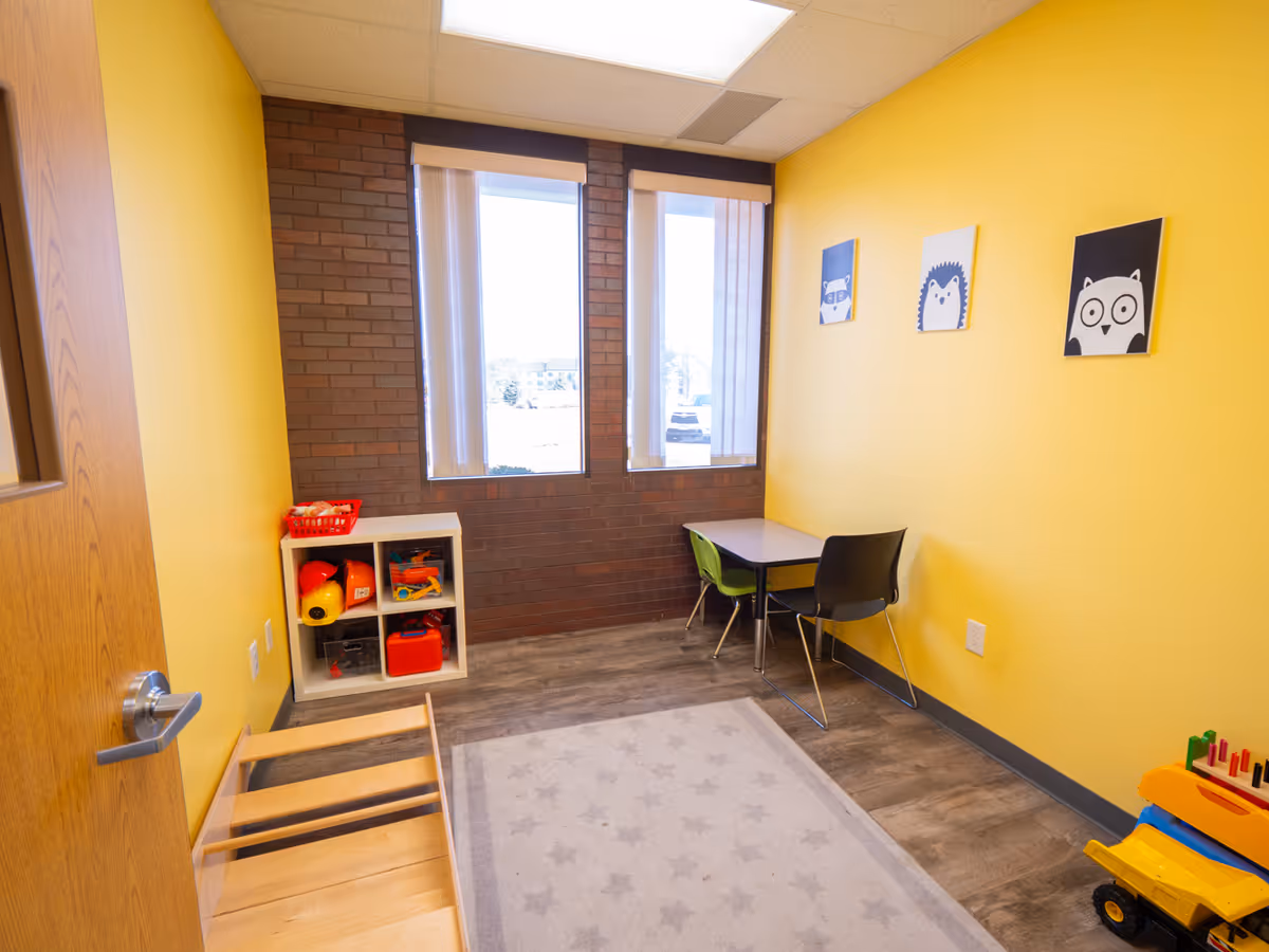 Yellow playroom with toy shelf, small table and chairs, starry rug, and windows.