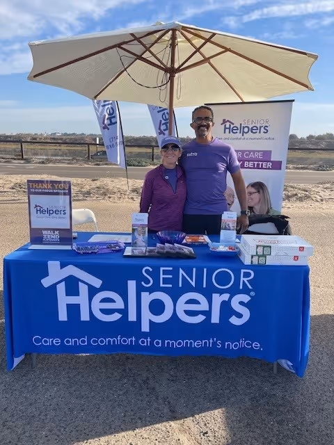 Two staff members stand behind a blue Senior Helpers booth table under a large umbrella outdoors.