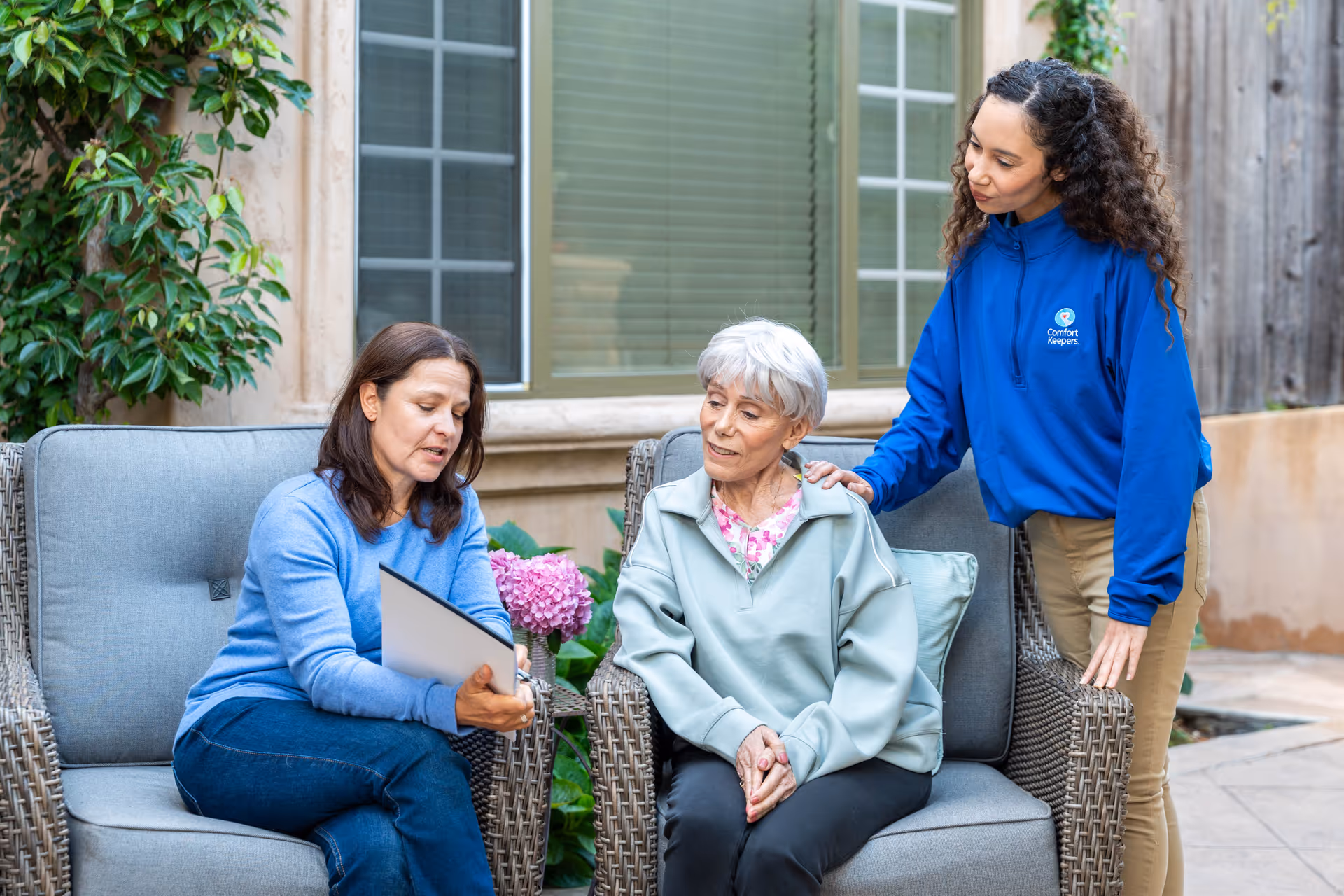 Three people converse outdoors on a patio; a Comfort Keepers caregiver assists an elderly woman.