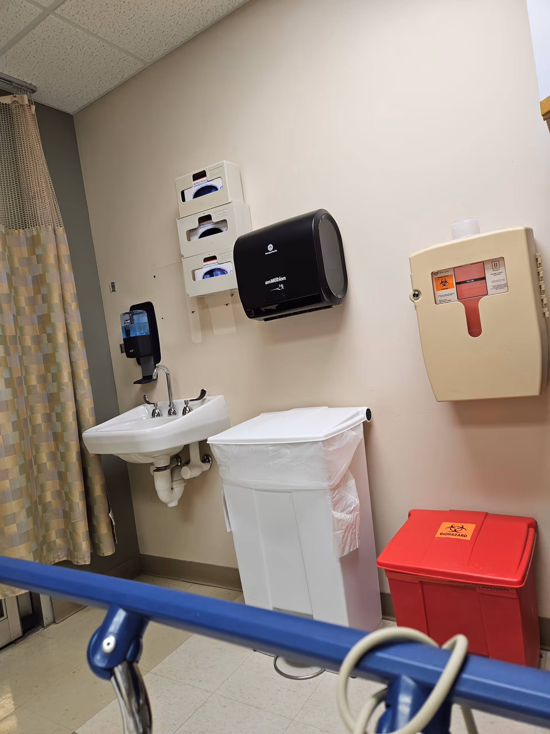 Medical room corner with sink, paper towels, sharps box, red biohazard bin and curtain