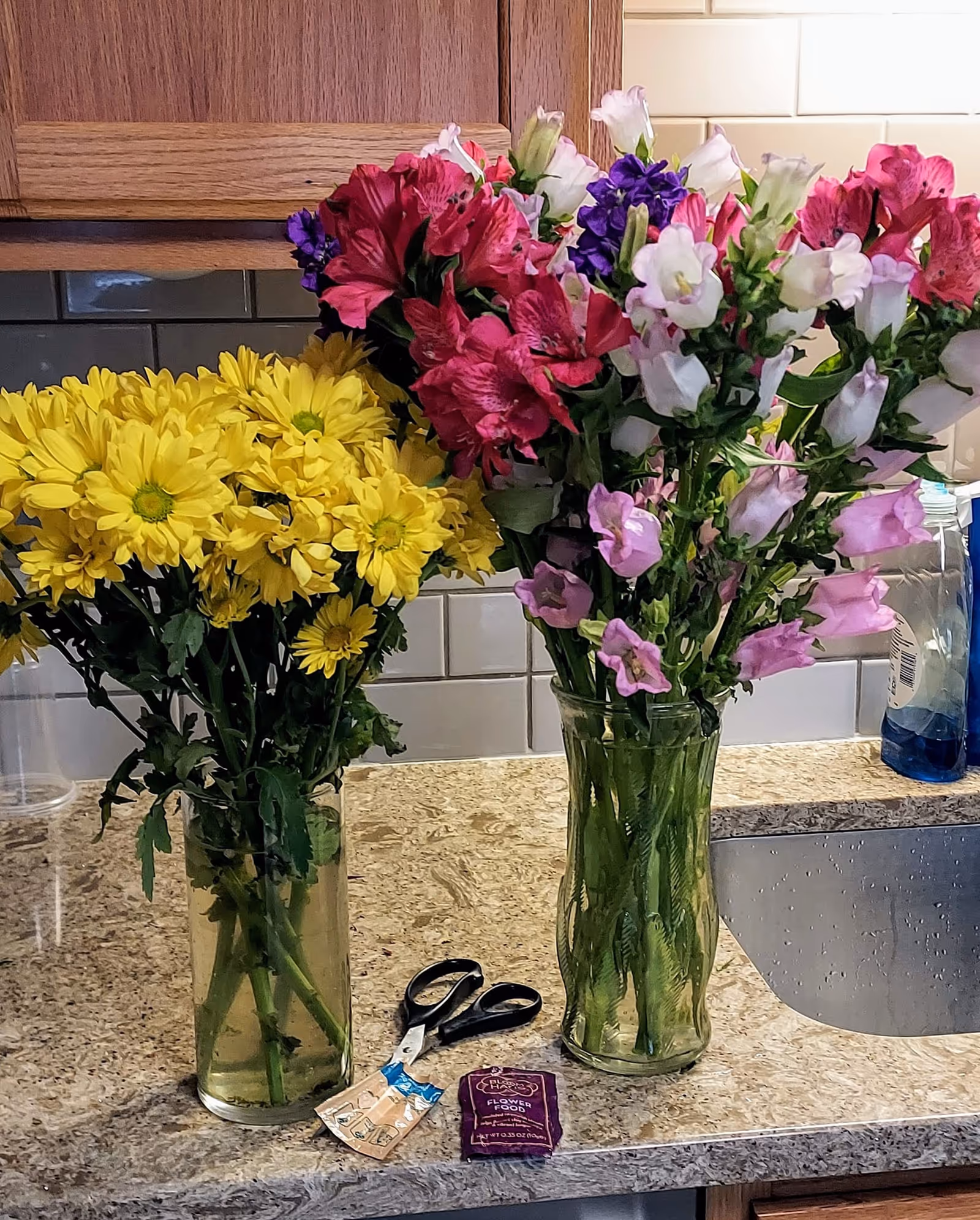 Two glass vases of colorful flowers sit on a marble kitchen counter with scissors nearby.