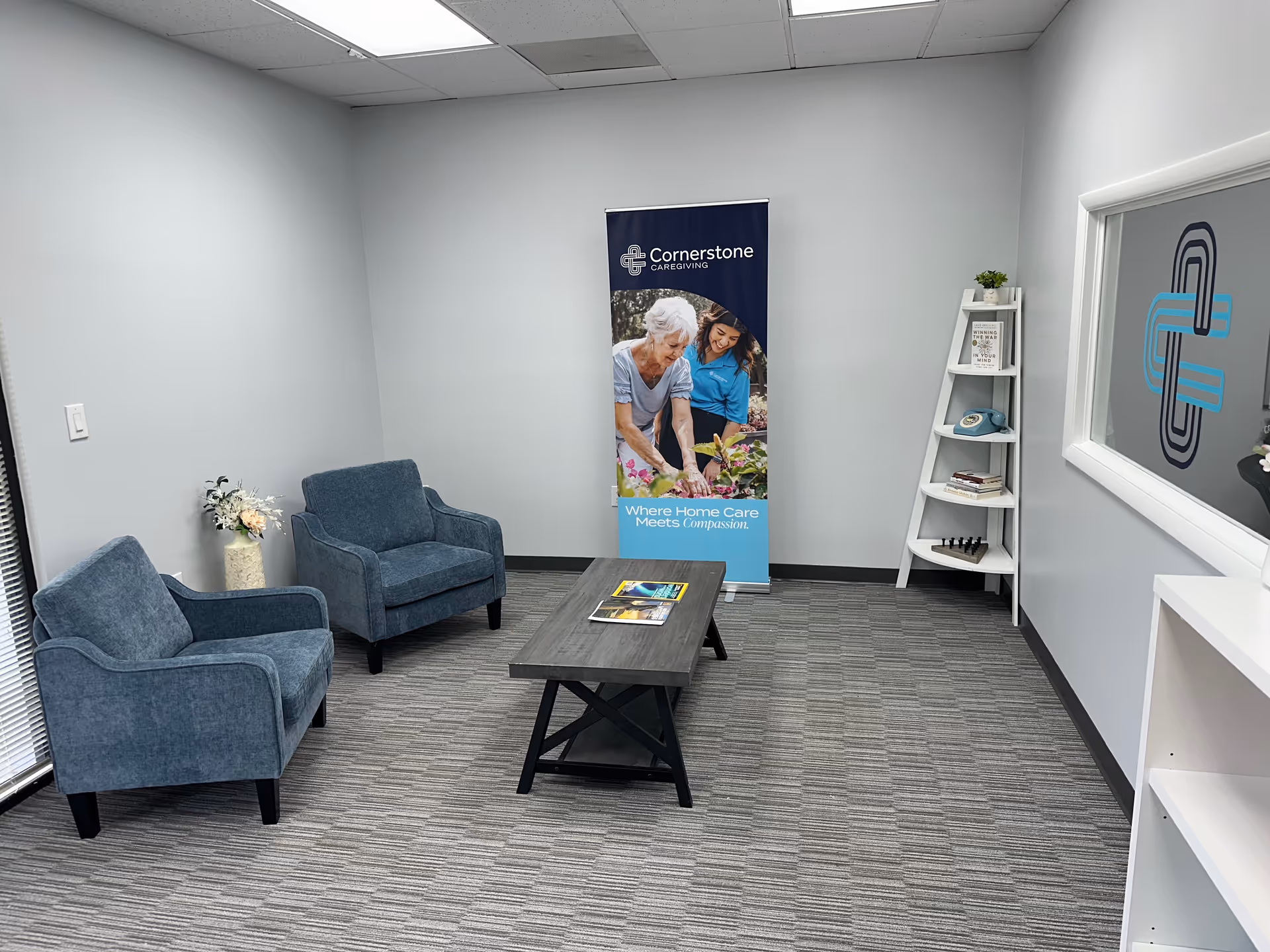 Interior waiting area with two blue chairs, a banner, coffee table, and a white bookshelf.