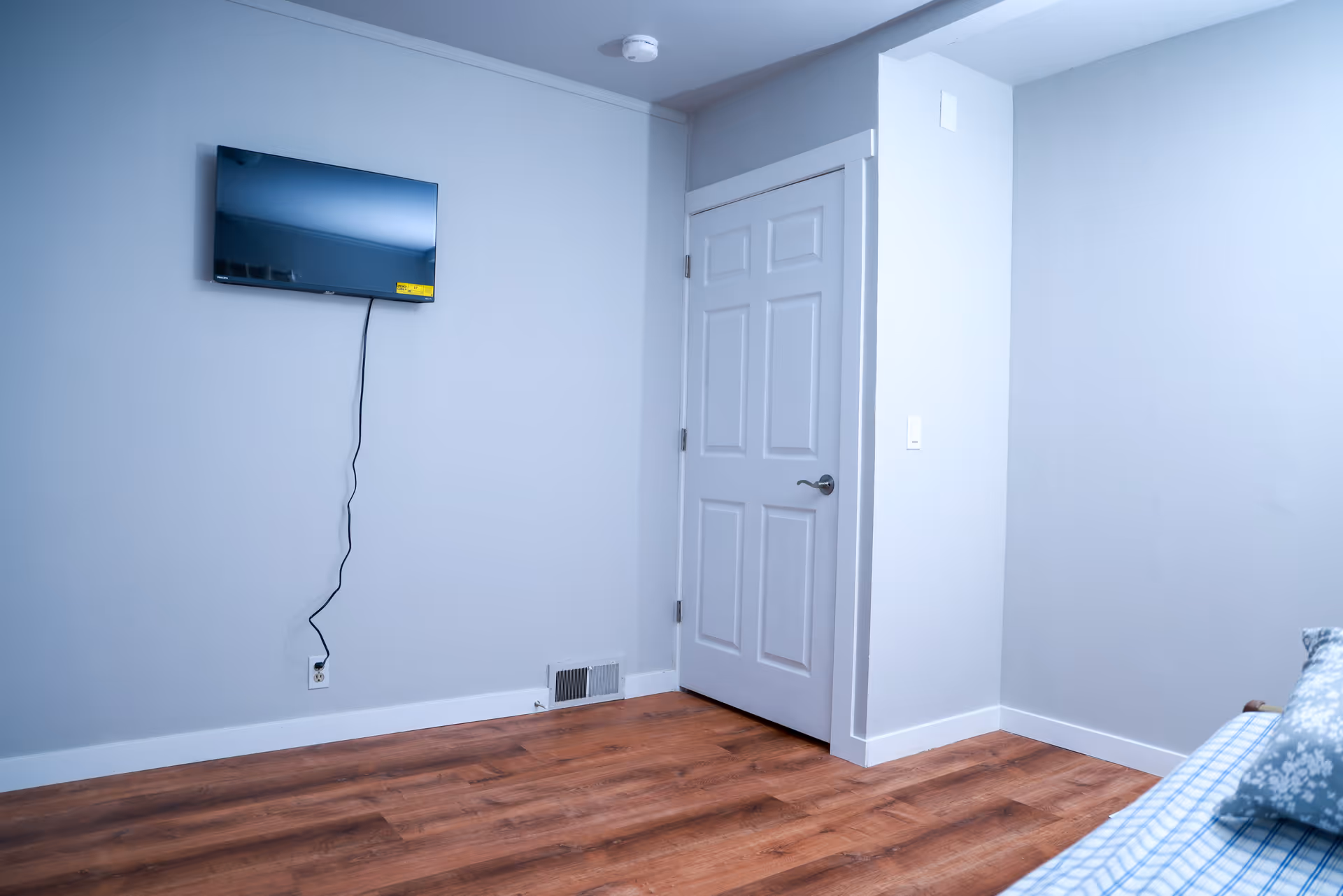 Sparsely furnished bedroom with a wall-mounted TV, wood floor, and a closed white door.