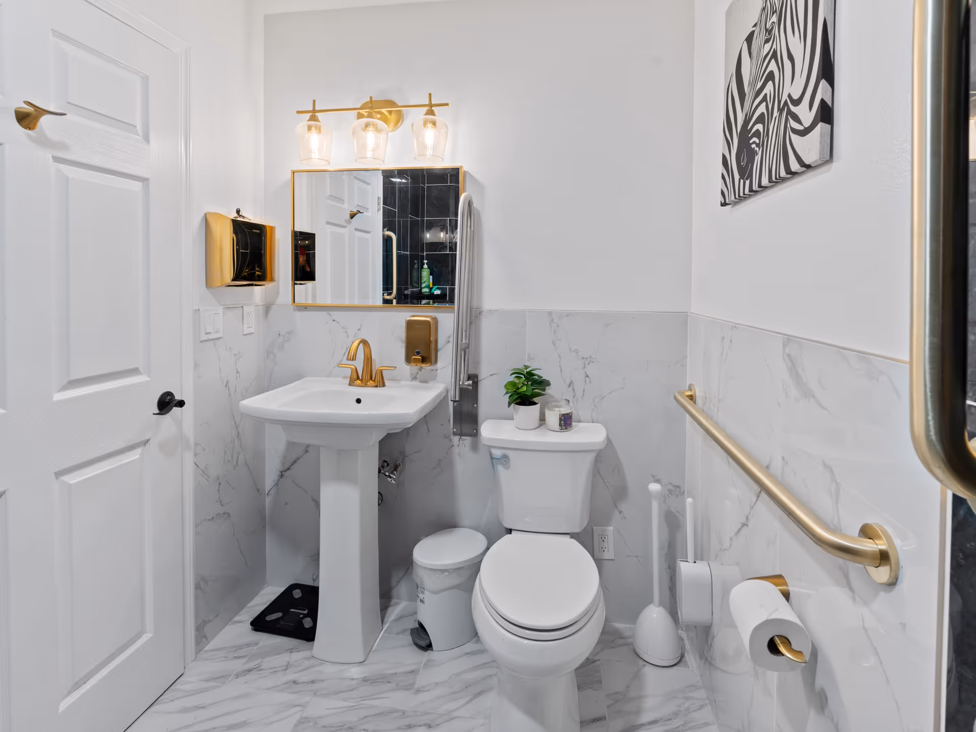 Bright marble-tiled bathroom with pedestal sink, toilet, gold fixtures, and a wall grab bar.