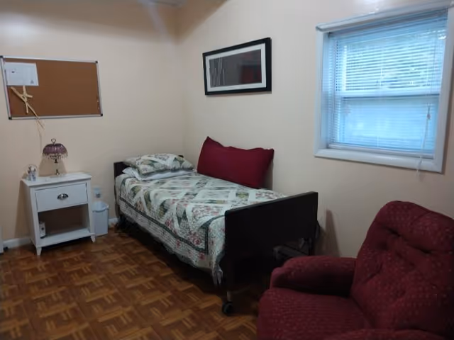 A small bedroom in a senior living facility with a bed, nightstand, and red chair.