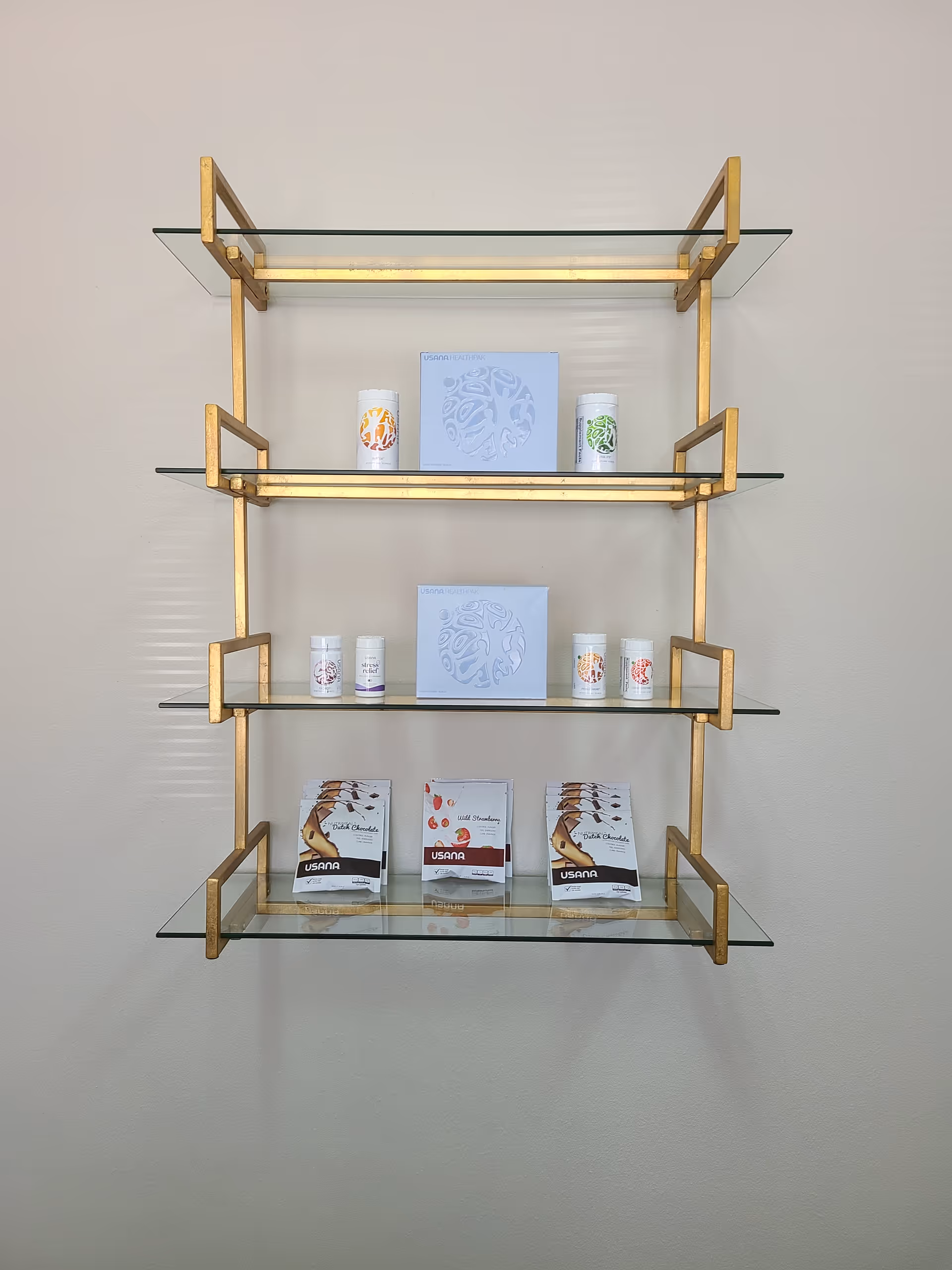 Wall-mounted glass shelving unit with gold brackets displaying cosmetics and chocolate bars.