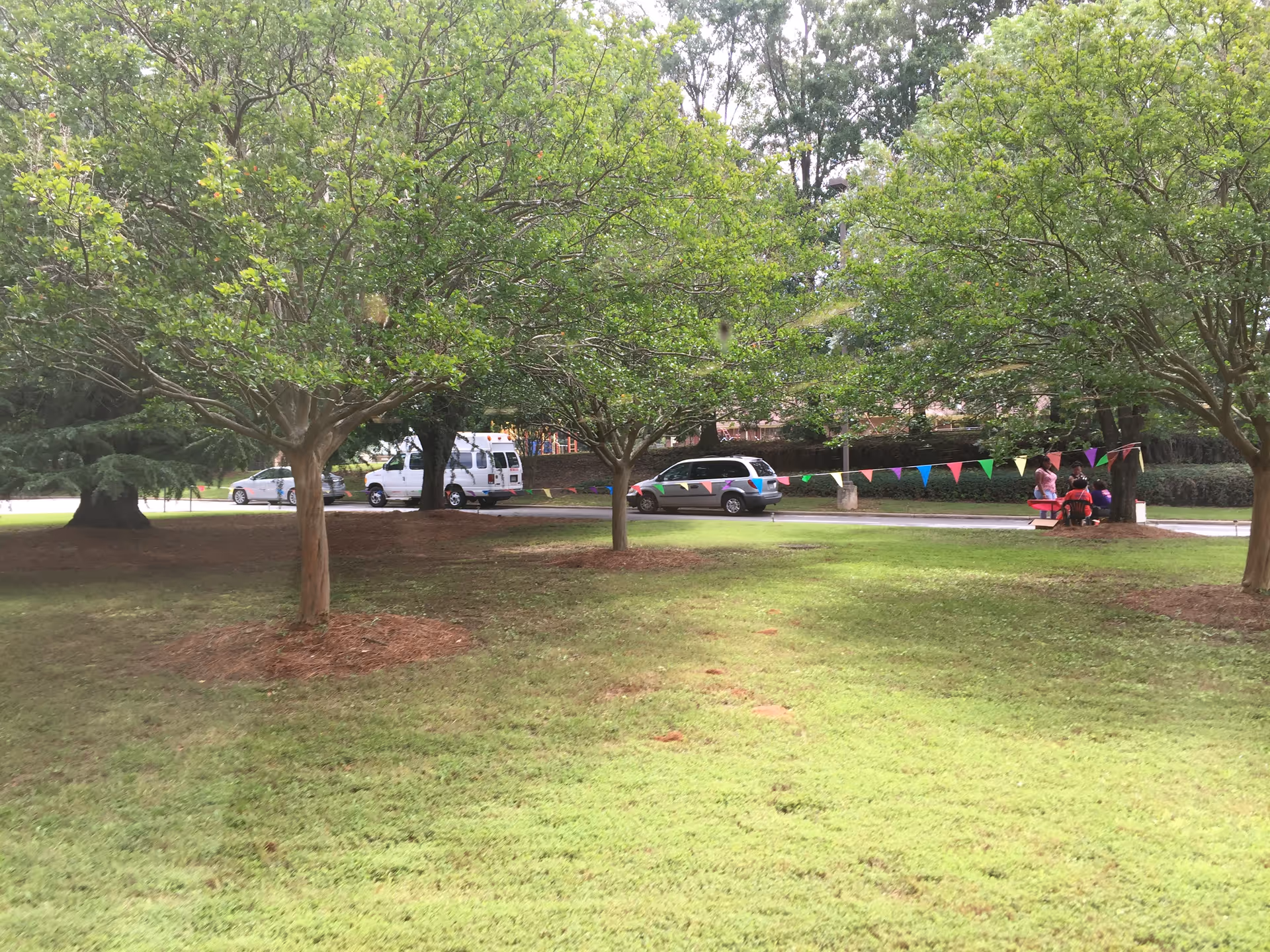 Outdoor park area with trees, bunting, vehicles, and a table with seating.