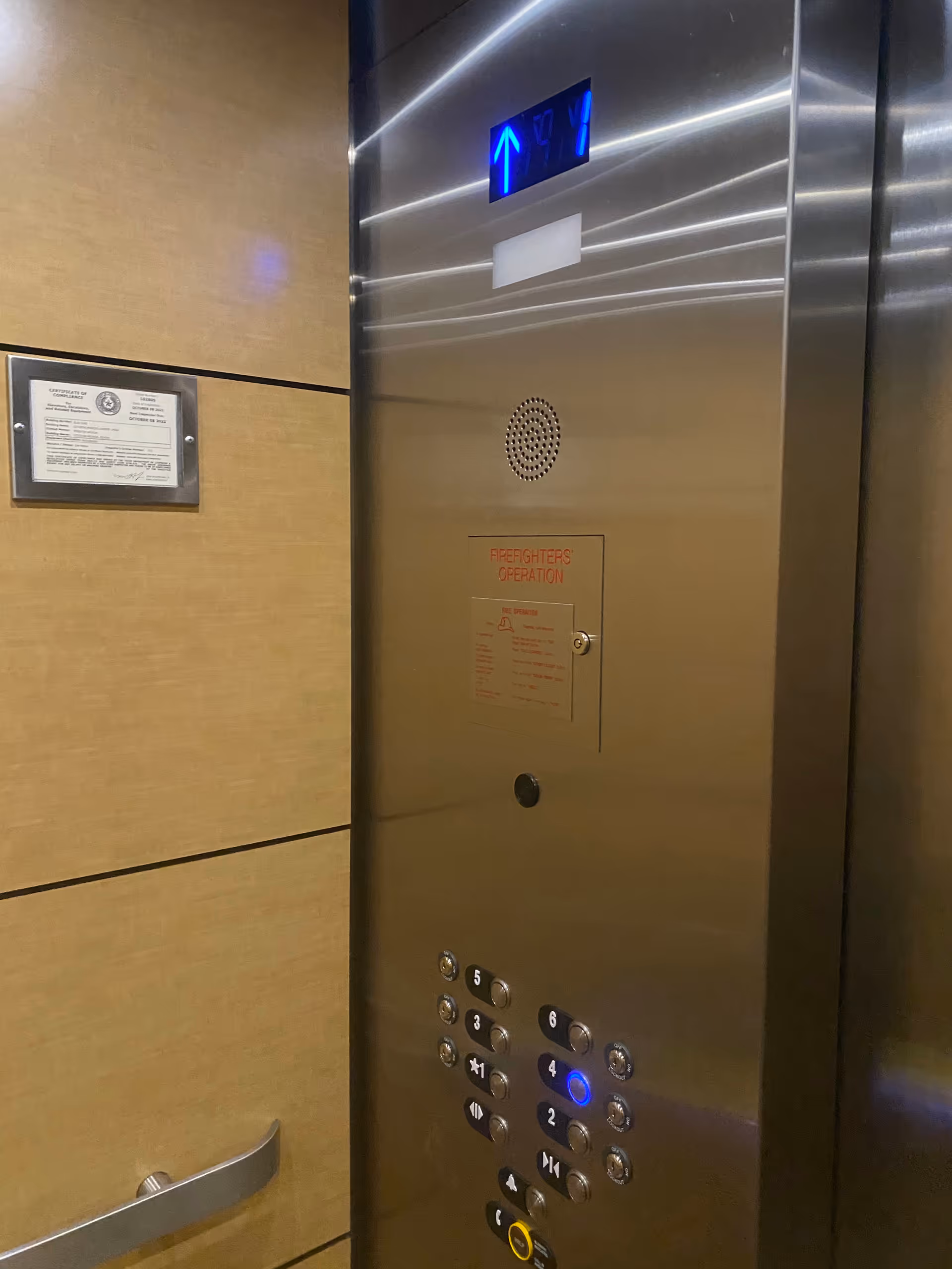 A stainless steel elevator interior with control panel, blue arrow display, and wooden wall panels.