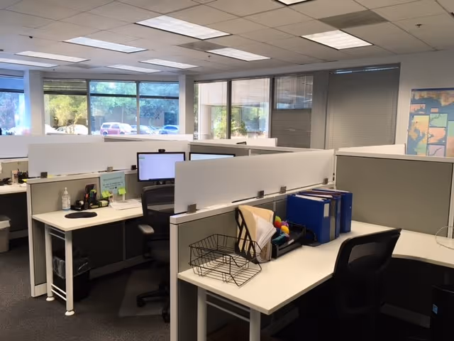 Open-plan office with cubicles, desks, computers, and partition panels in a bright space.
