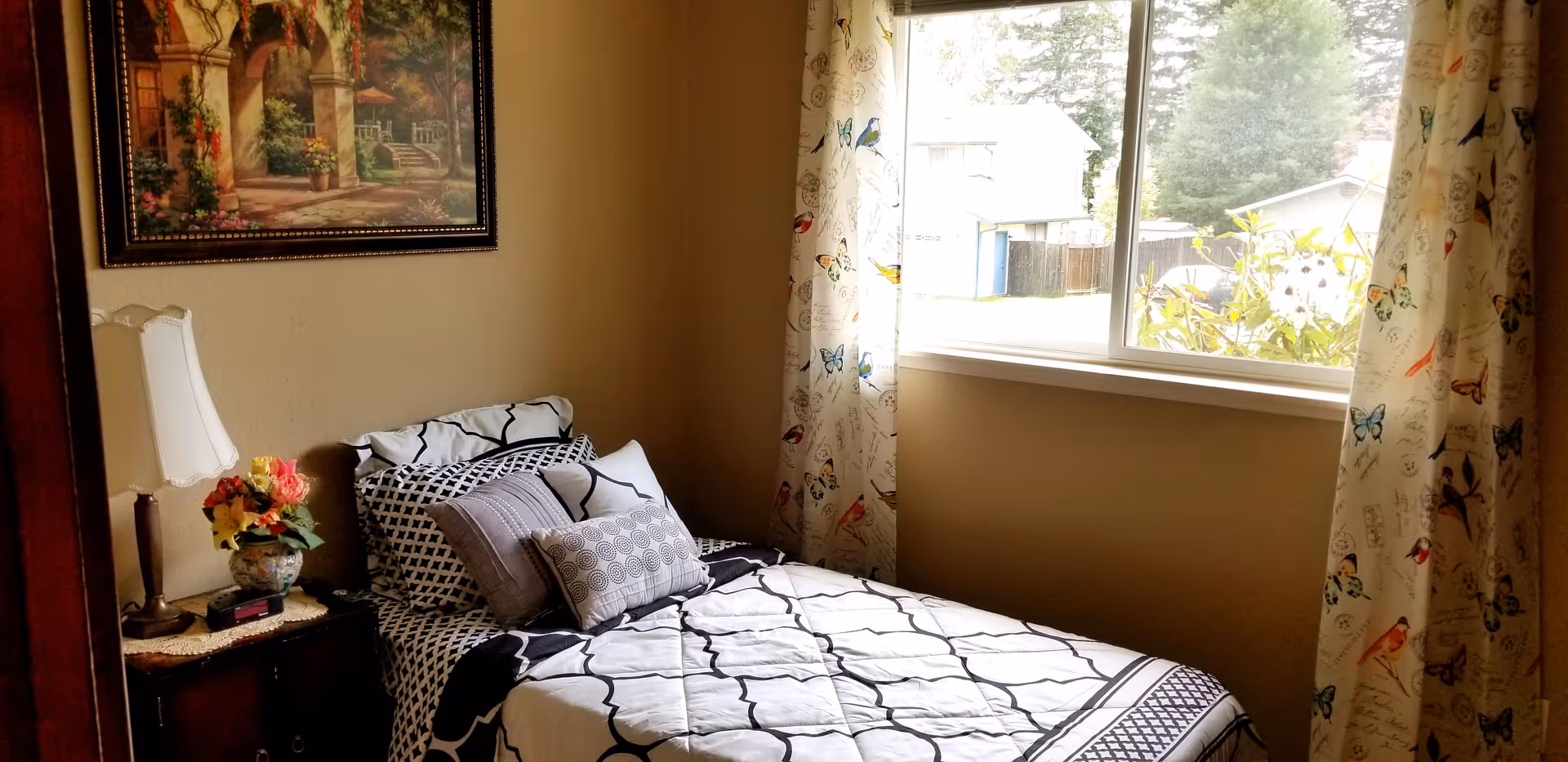 Cozy bedroom with a bed, nightstand lamp, and butterfly-pattern curtains near a window.