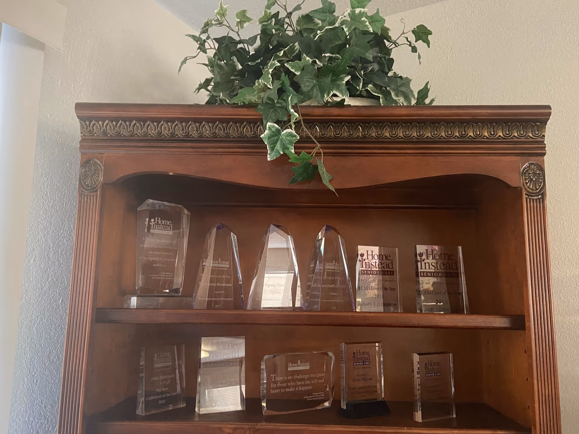 Wood display cabinet with multiple Home Instead awards and a potted plant on top.