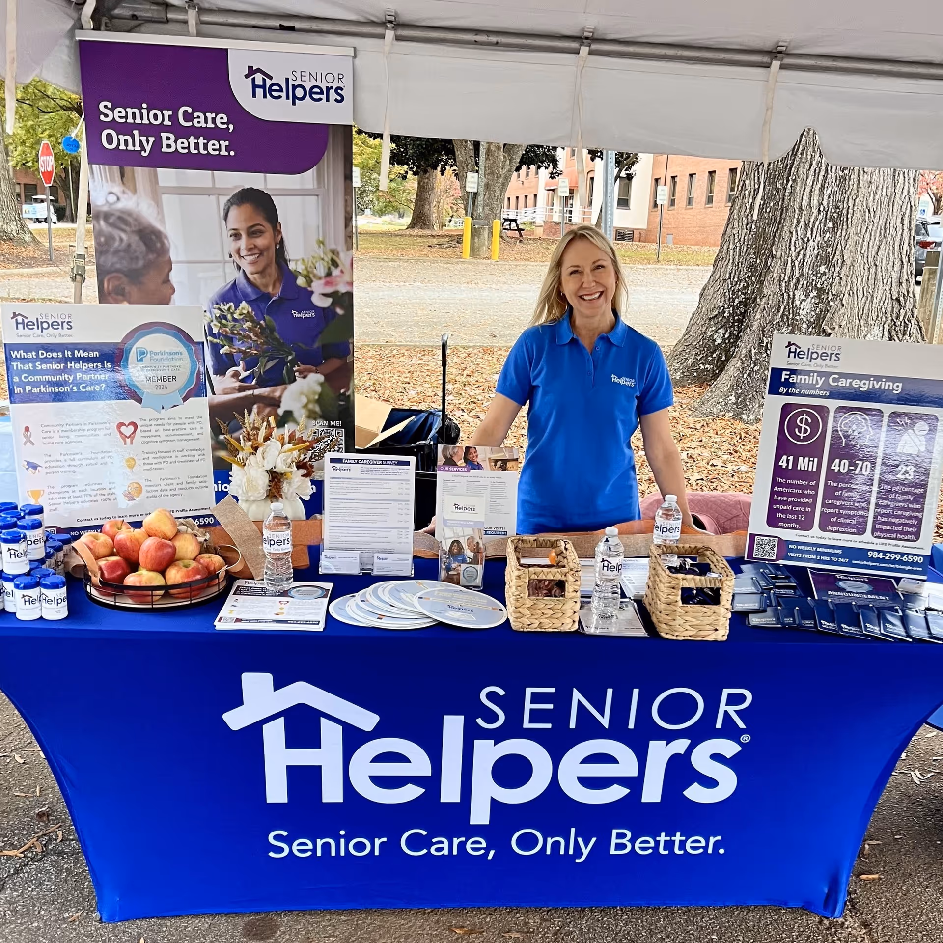 Smiling staff member at Senior Helpers booth outdoors under a canopy.