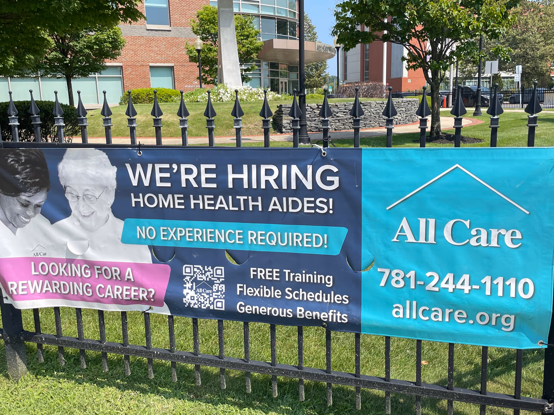 Banner on a black fence advertising All Care hiring in front of a brick building.