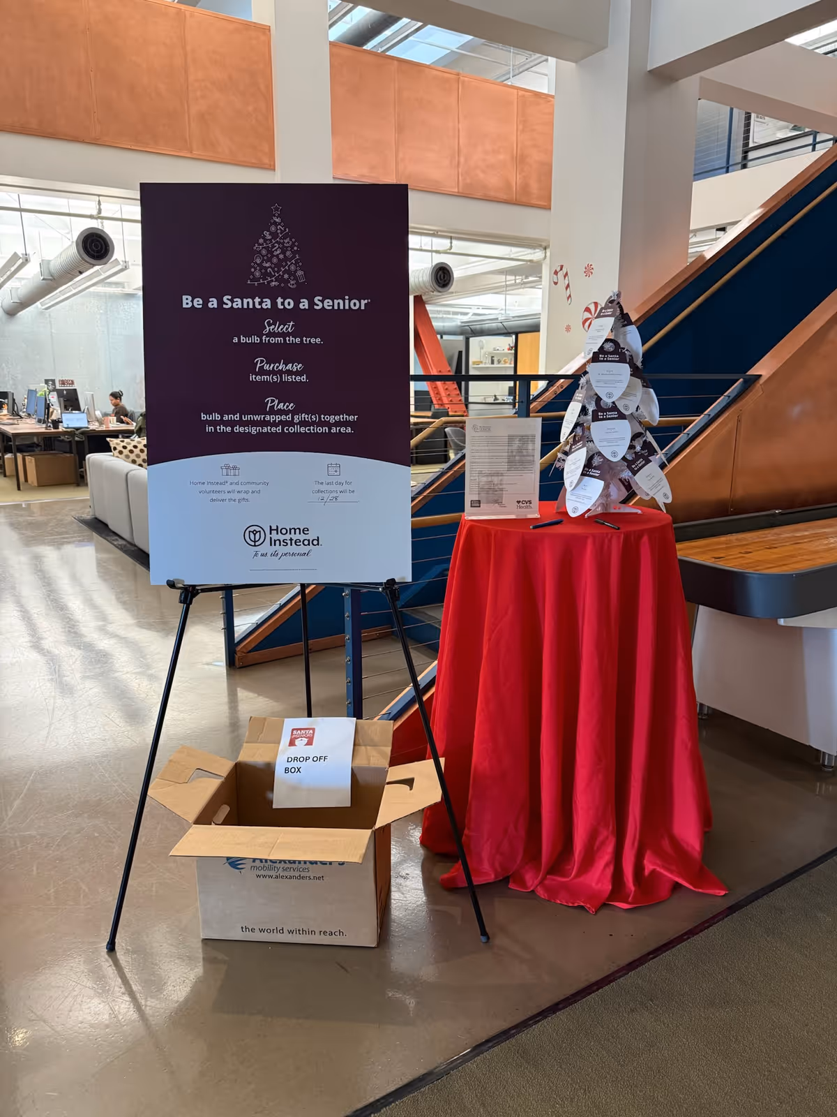 Indoor lobby with Be a Santa to a Senior sign, donation box, and red table cover.
