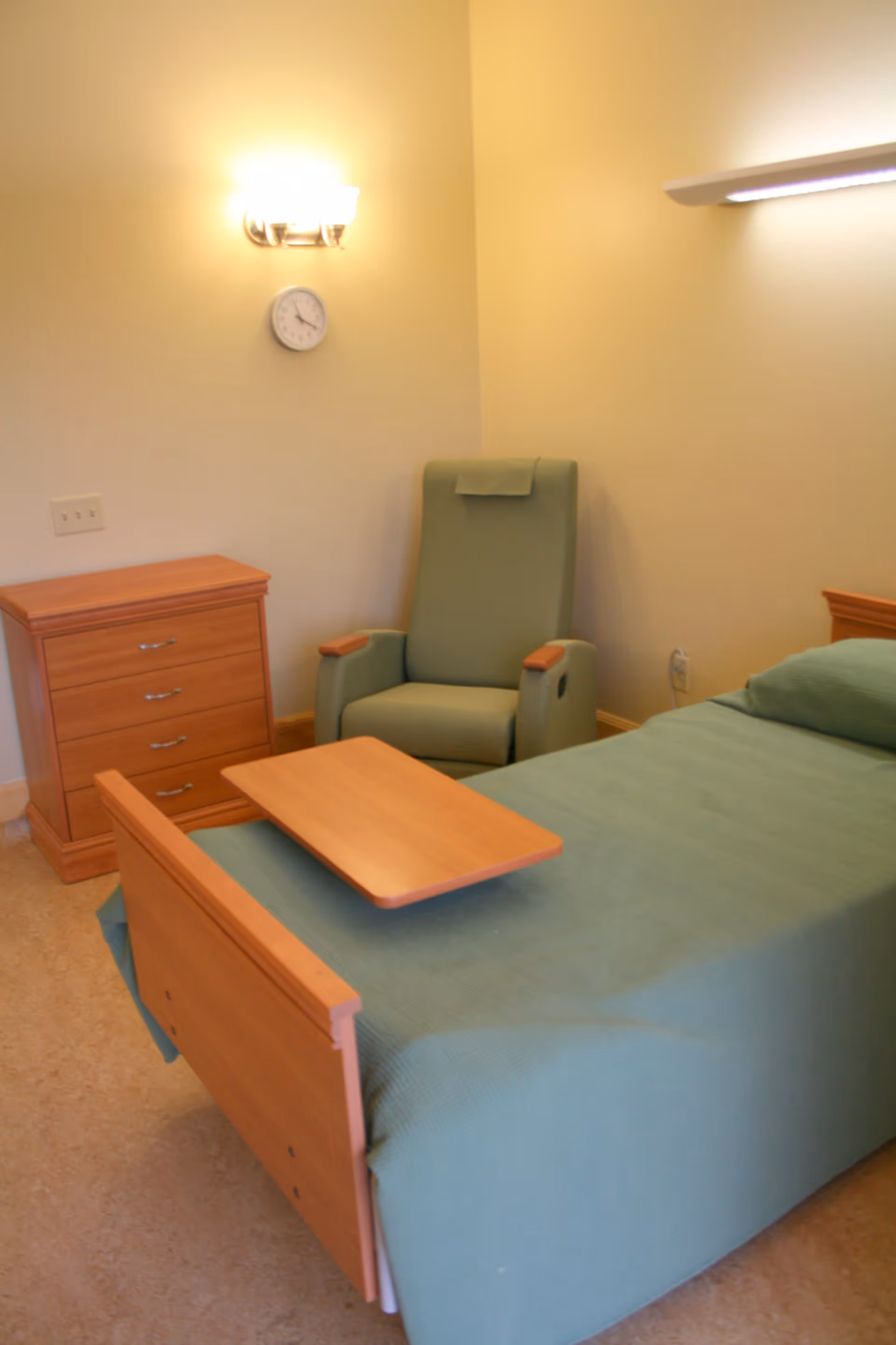 Hospice bedroom with green bed, chair, and wooden dresser in soft lighting.