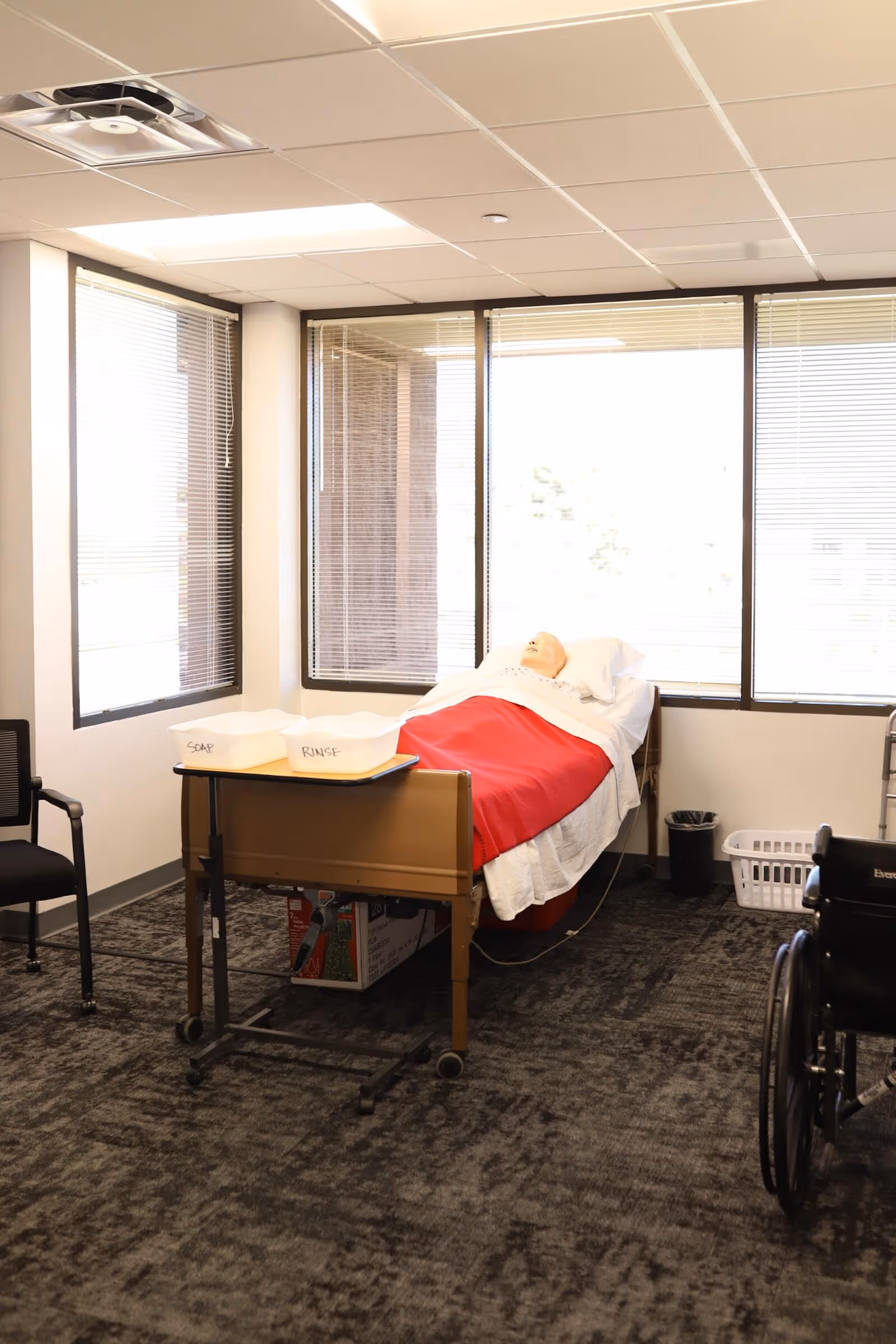Hospital bed with red blanket and pillow, wheelchair nearby in a bright interior room.