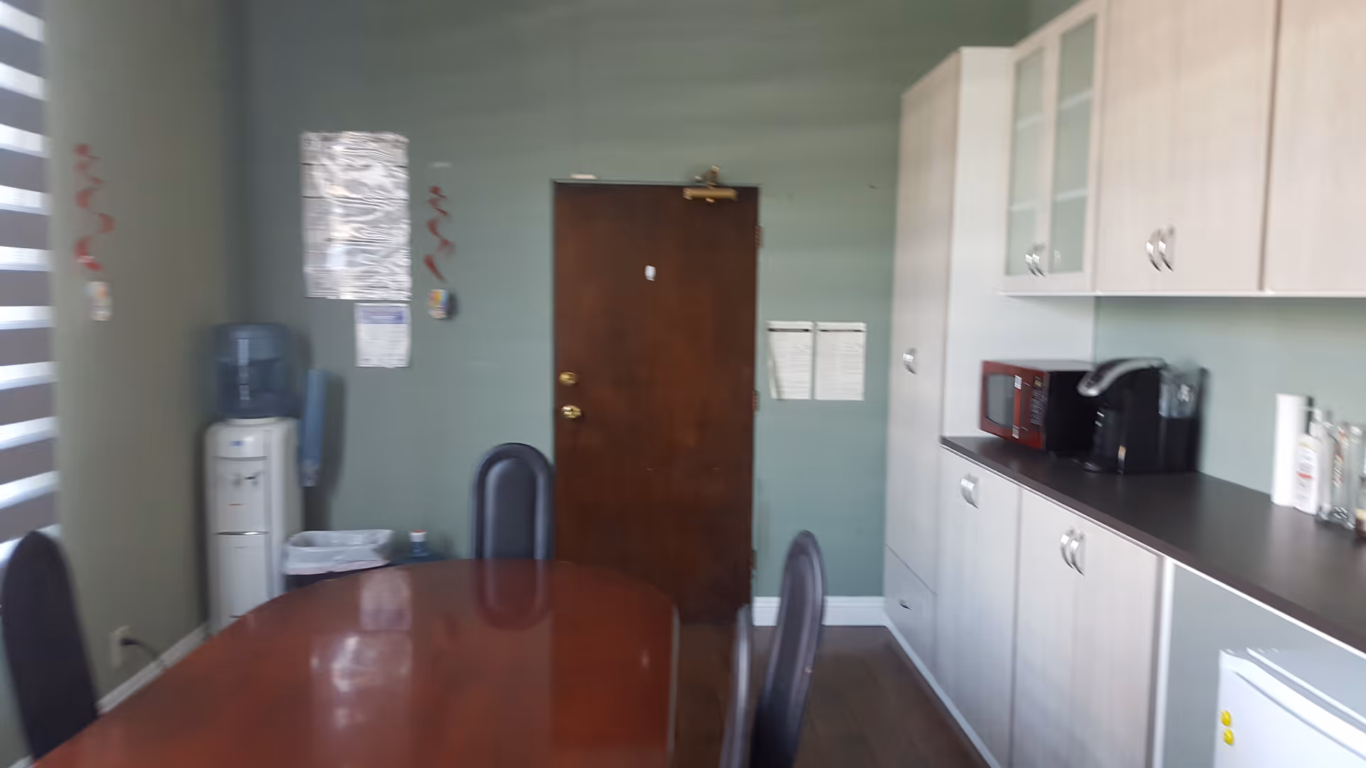 A senior living facility dining area with a long wooden table, chairs, and kitchenette along wall.