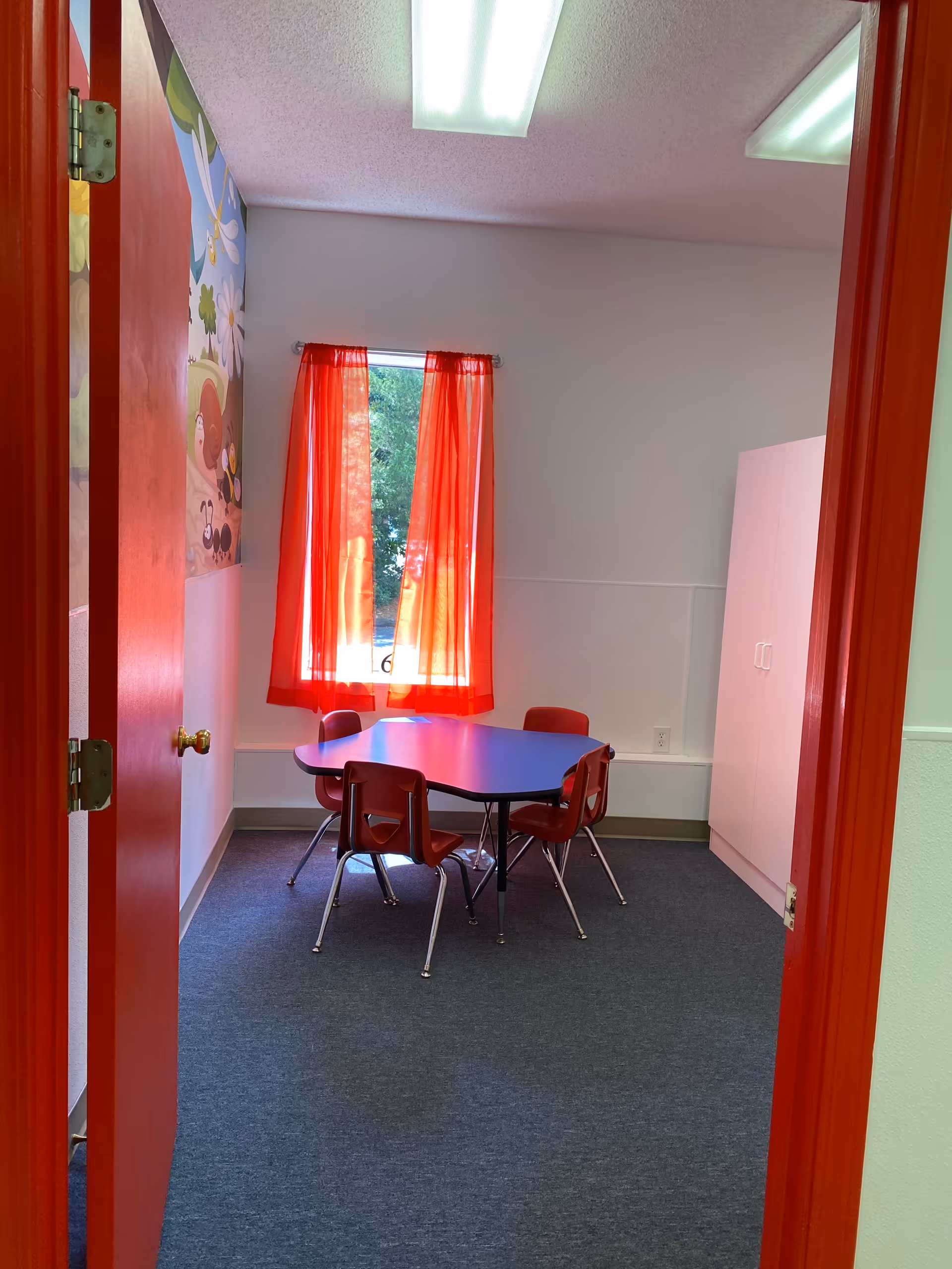 Small interior therapy room with red curtains, blue table, red chairs, and mural on the wall.