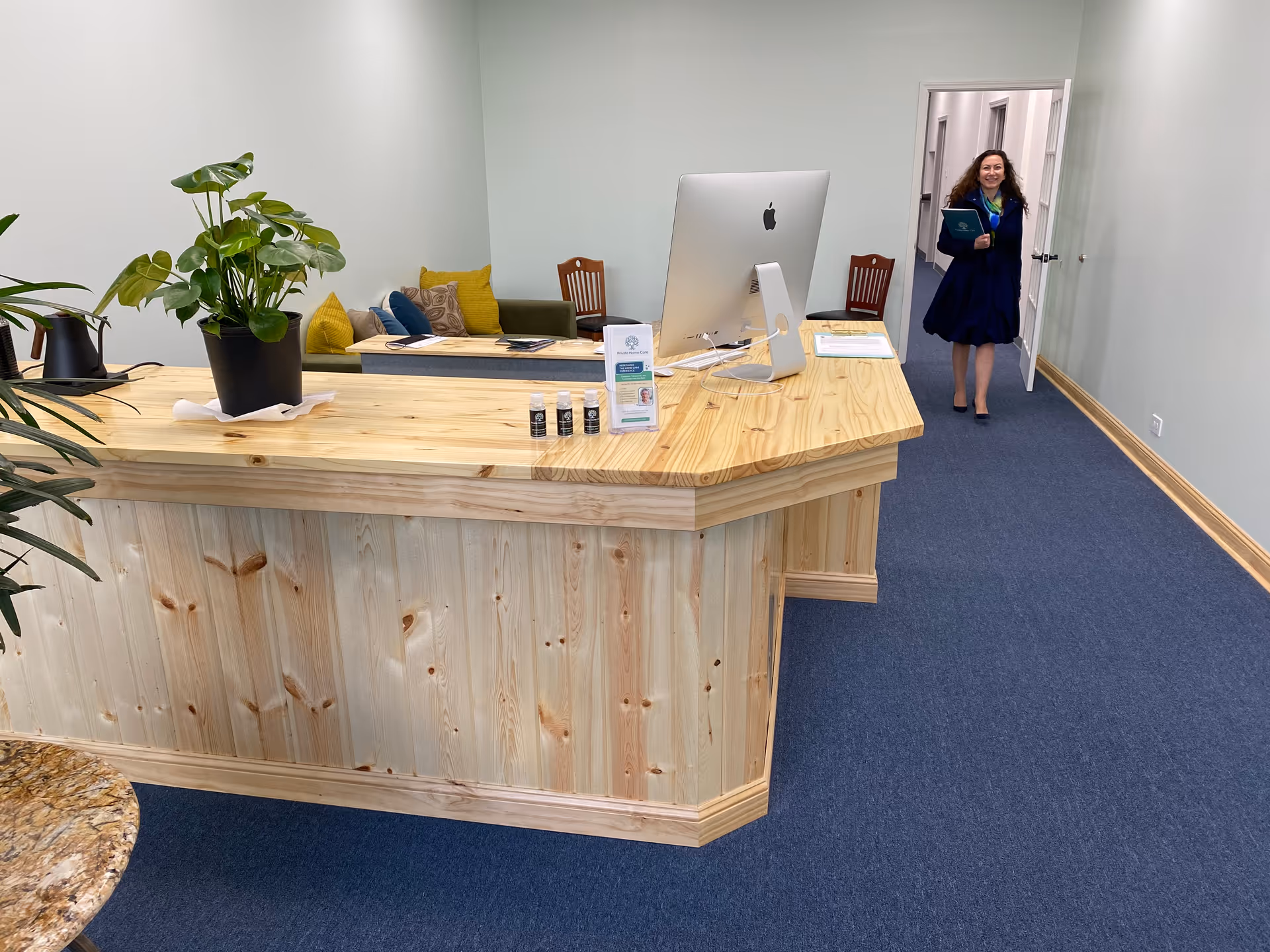Reception desk in a light wood lobby as a woman enters from the hallway.