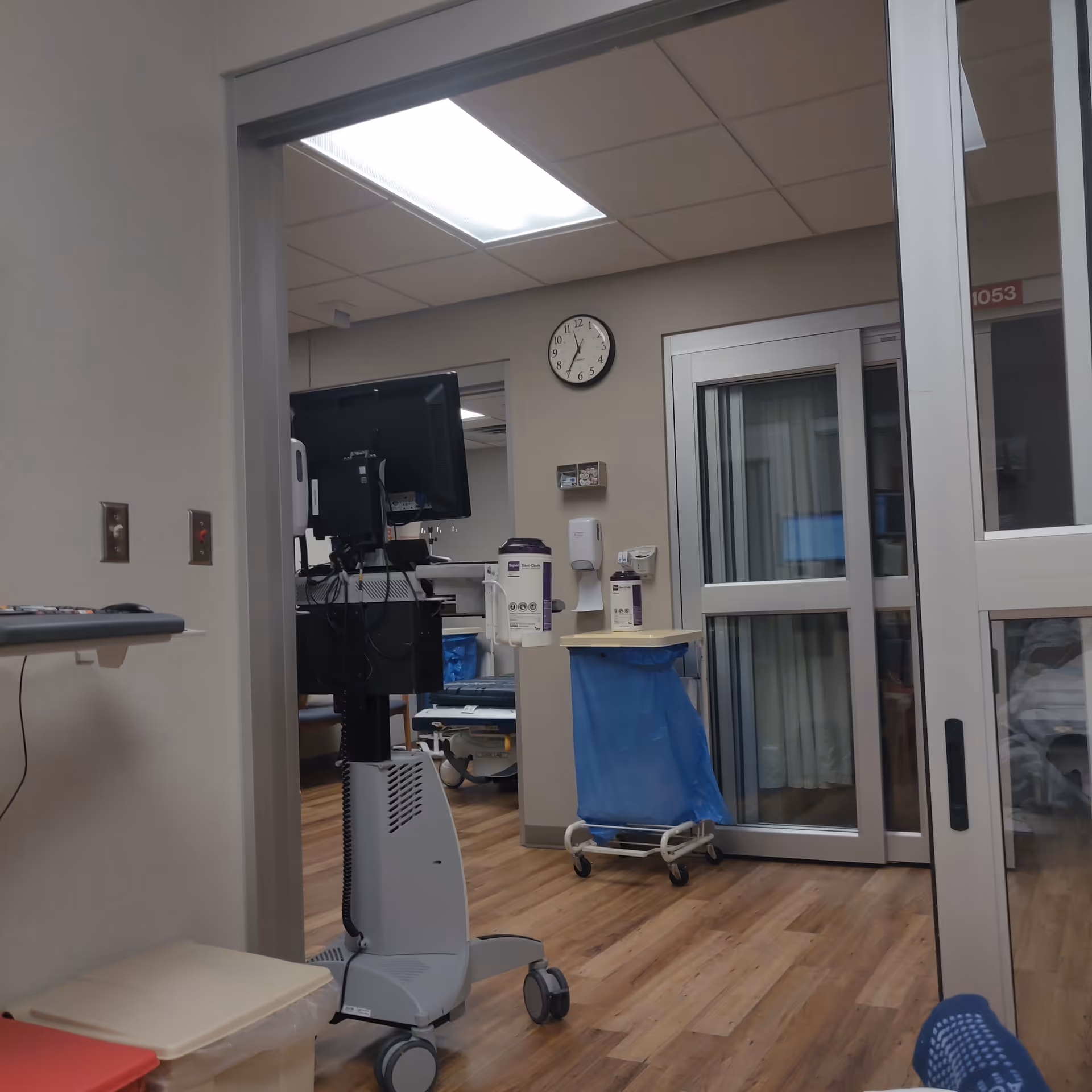 Hospital corridor with medical equipment, computer monitor, clock, and a blue waste cart.