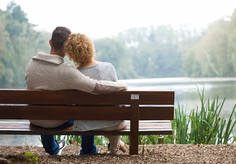 Two people sit on a wooden bench by a lake, embracing.
