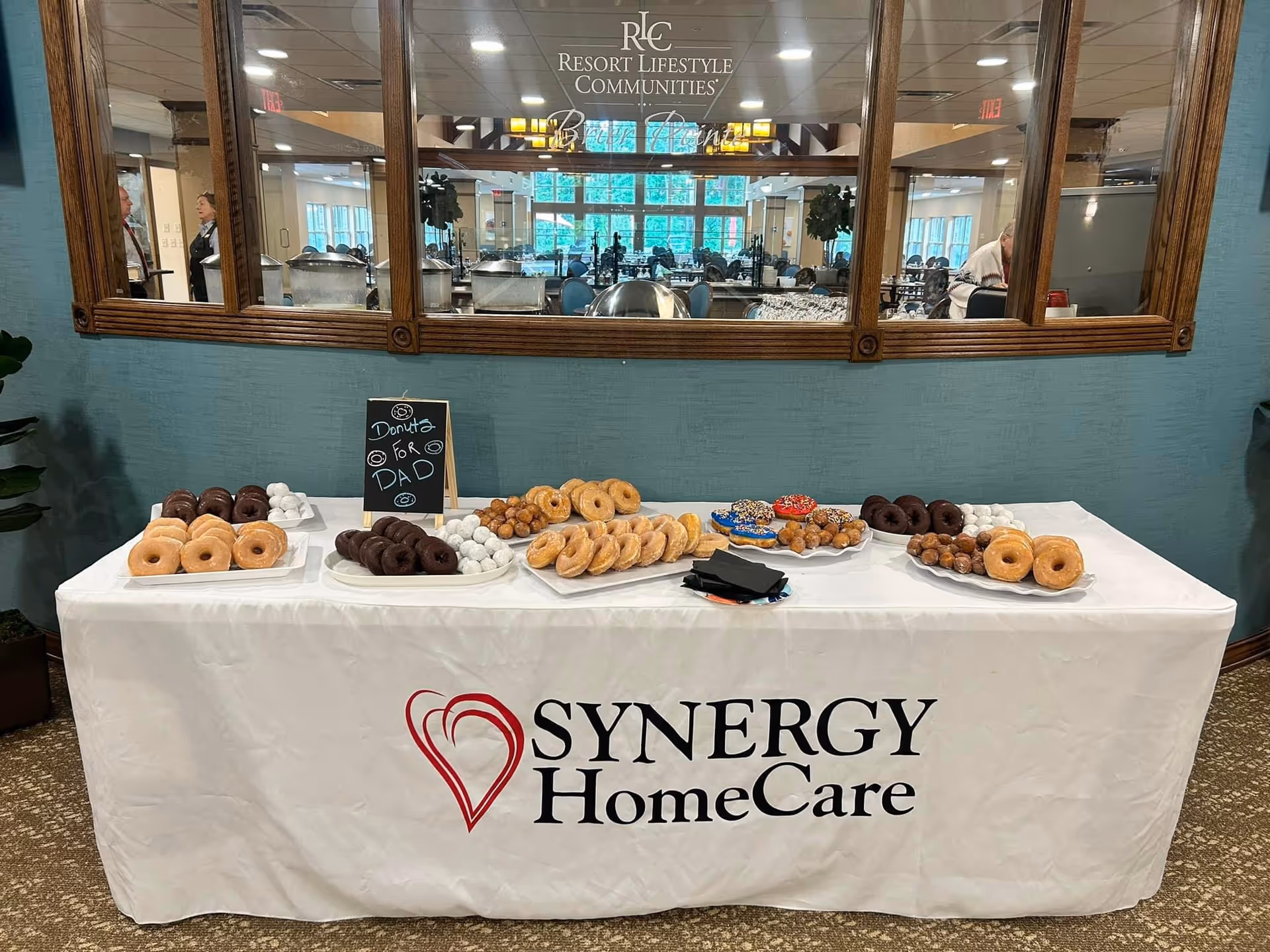 Donut and pastry table branded SYNERGY HomeCare in a facility lobby area.