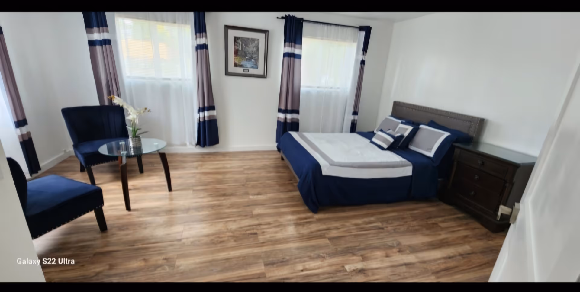 Blue-and-white themed bedroom with bed, two chairs, glass table, and nightstand.